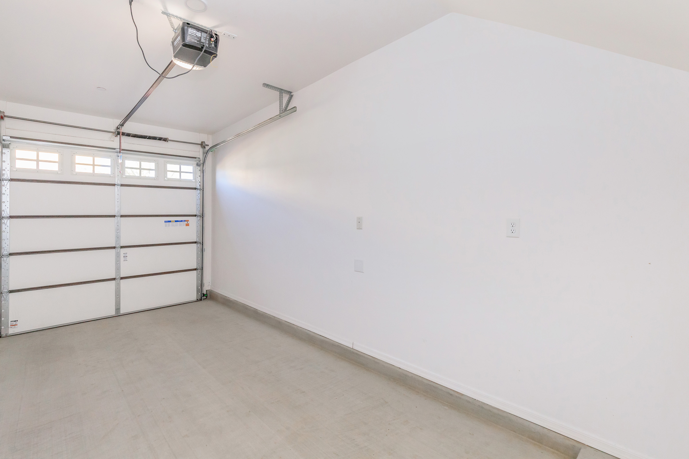 A clean, empty garage space with white walls and a concrete floor. The garage door is partially open, revealing natural light entering the room. There are no vehicles or storage items present, creating an unfinished and open atmosphere.