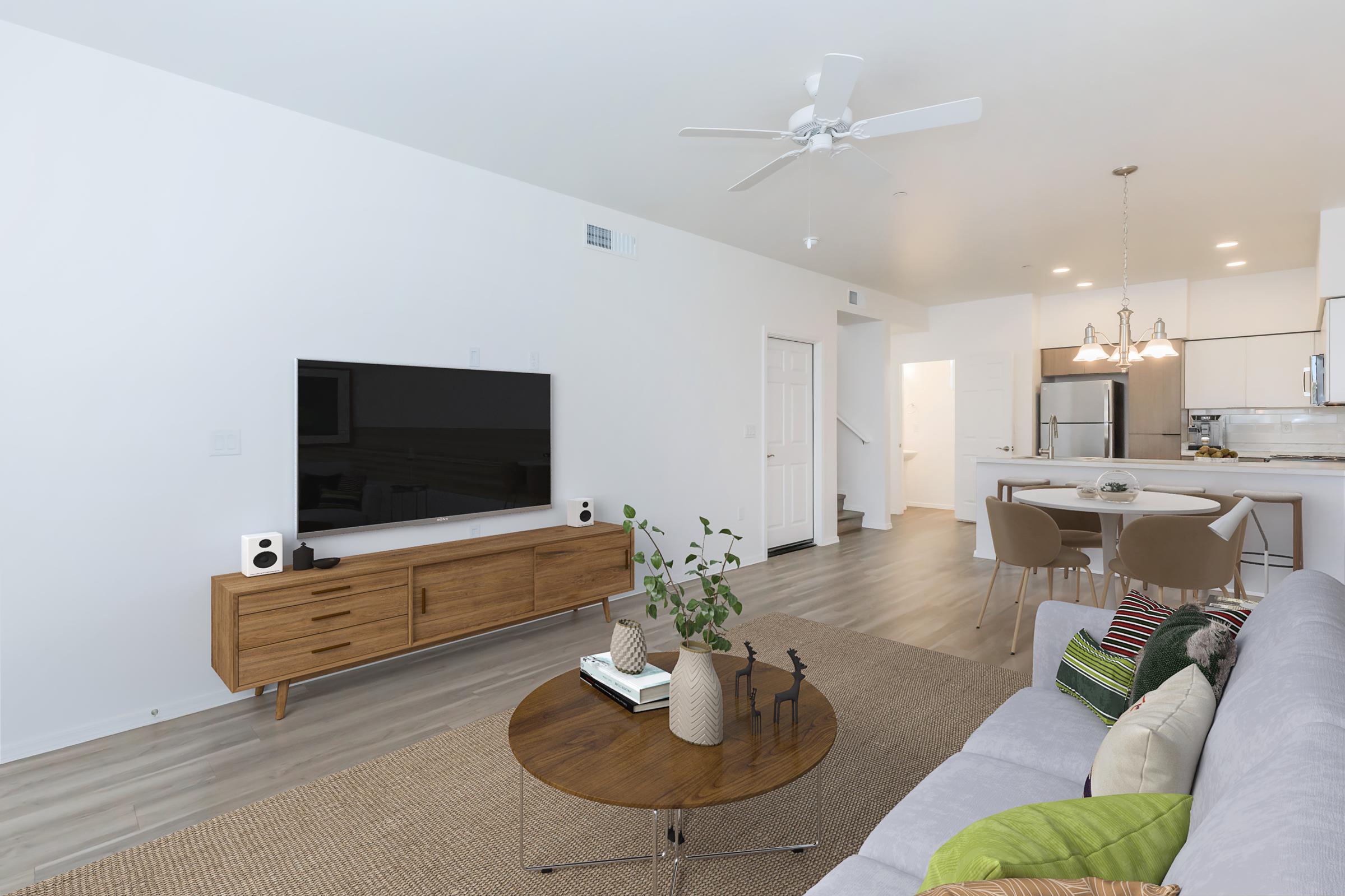 A modern living room featuring a gray sofa with colorful pillows, a circular wooden coffee table, and a large TV mounted on the wall. There is a wooden sideboard beneath the TV, and a kitchen area can be seen in the background with bar stools and contemporary lighting fixtures. A houseplant adds a touch of greenery.