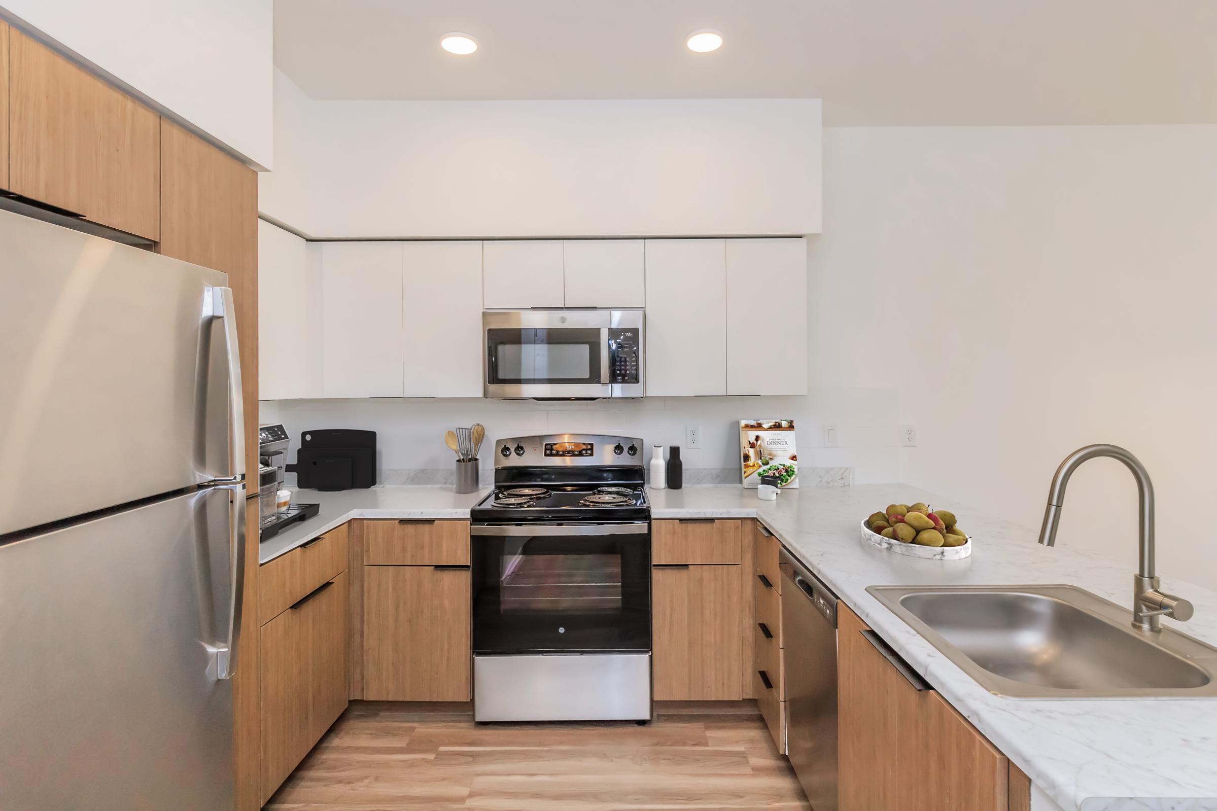 A modern kitchen featuring stainless steel appliances, including an oven, microwave, and refrigerator. The cabinetry is a mix of light wood and white, with a marble countertop. A sink is positioned near the counter, and a bowl of green fruit is displayed on the countertop, creating a stylish and functional space.
