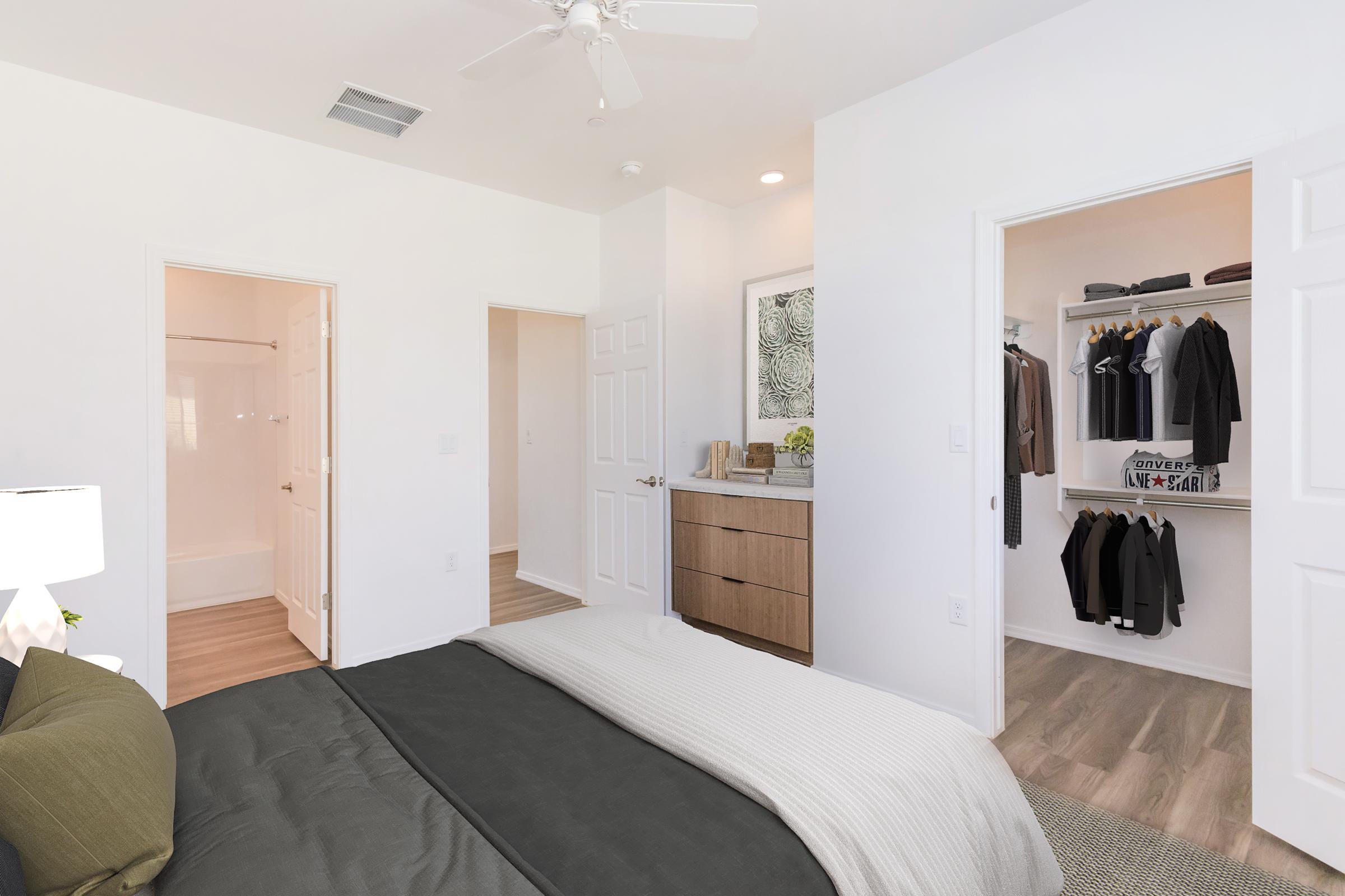 A modern bedroom featuring a large bed with dark bedding, a bedside lamp, and a stylish wall decor. Two doors lead to a bathroom and a walk-in closet filled with organized clothing. The room has light-colored walls and wooden flooring, creating a bright and spacious atmosphere.