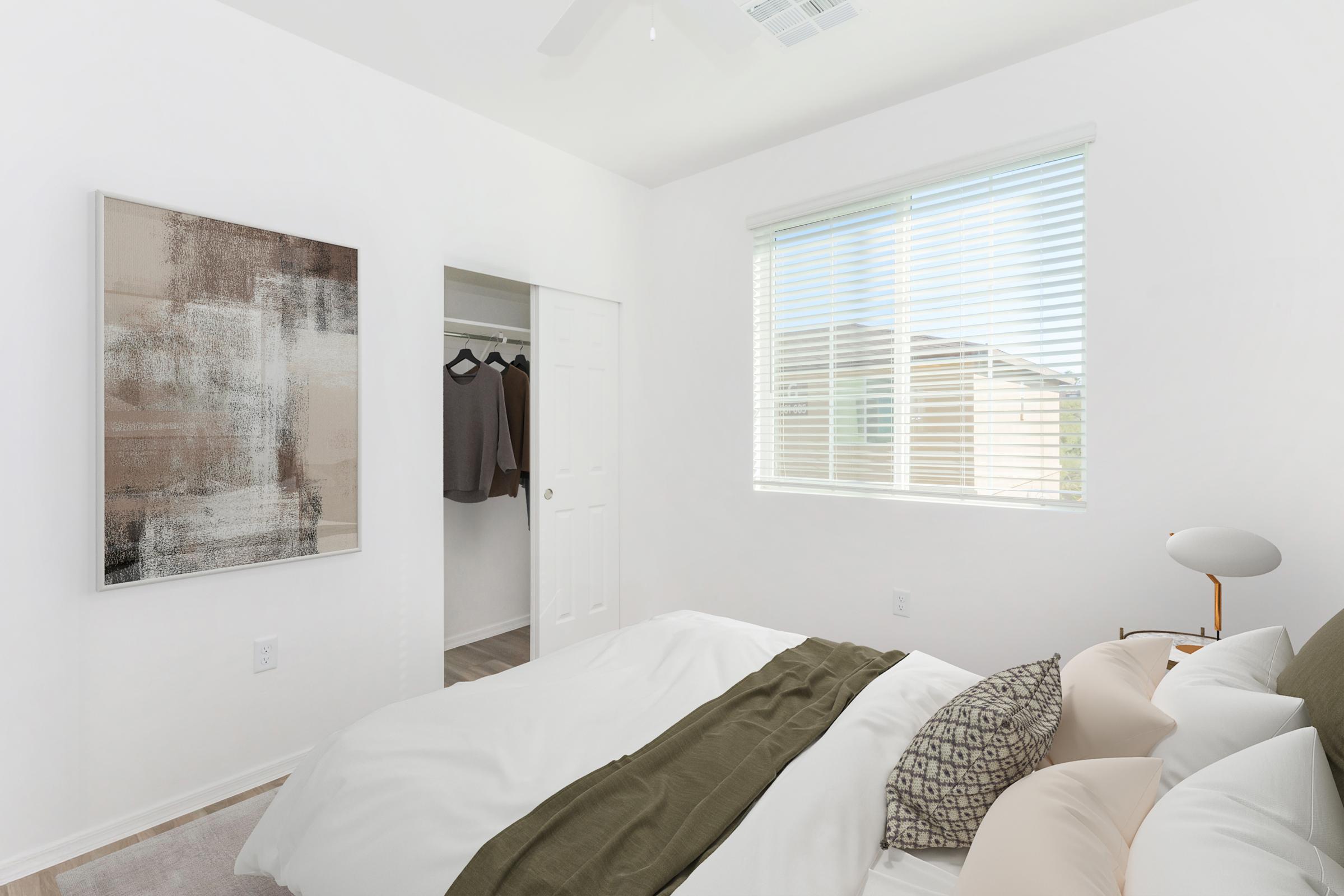 A bright and airy bedroom featuring a neatly made bed with white linens and decorative pillows. A modern artwork hangs on the wall beside a closet, which is partially open, revealing some clothing. Natural light streams in through a window with blinds, creating a welcoming atmosphere.