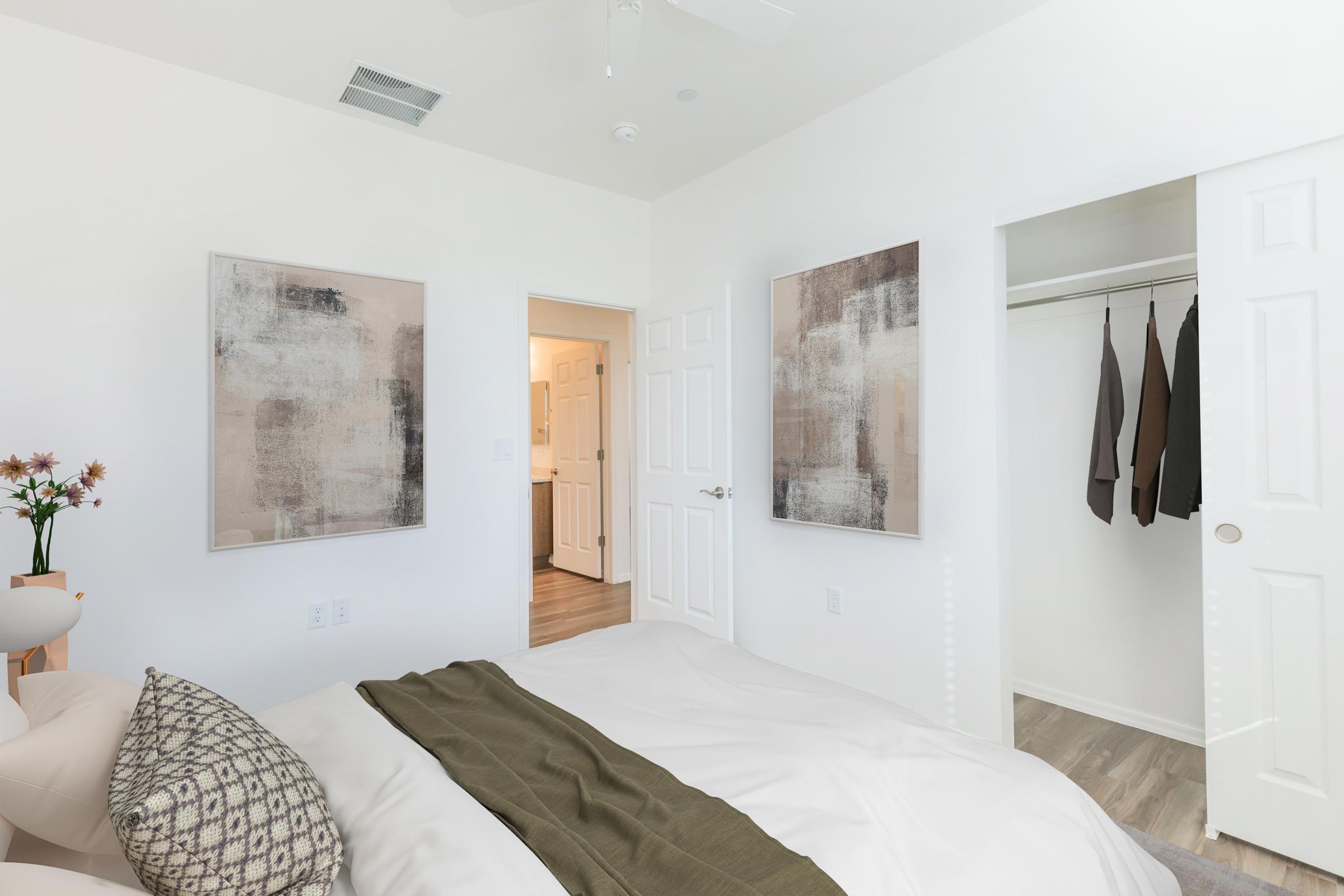 A bright, modern bedroom featuring a neatly made bed with a white comforter and decorative pillows. Two abstract art pieces hang on the walls, and a closet with sliding doors is partially visible. A doorway leading to a bathroom can be seen in the background, creating an inviting and airy atmosphere.