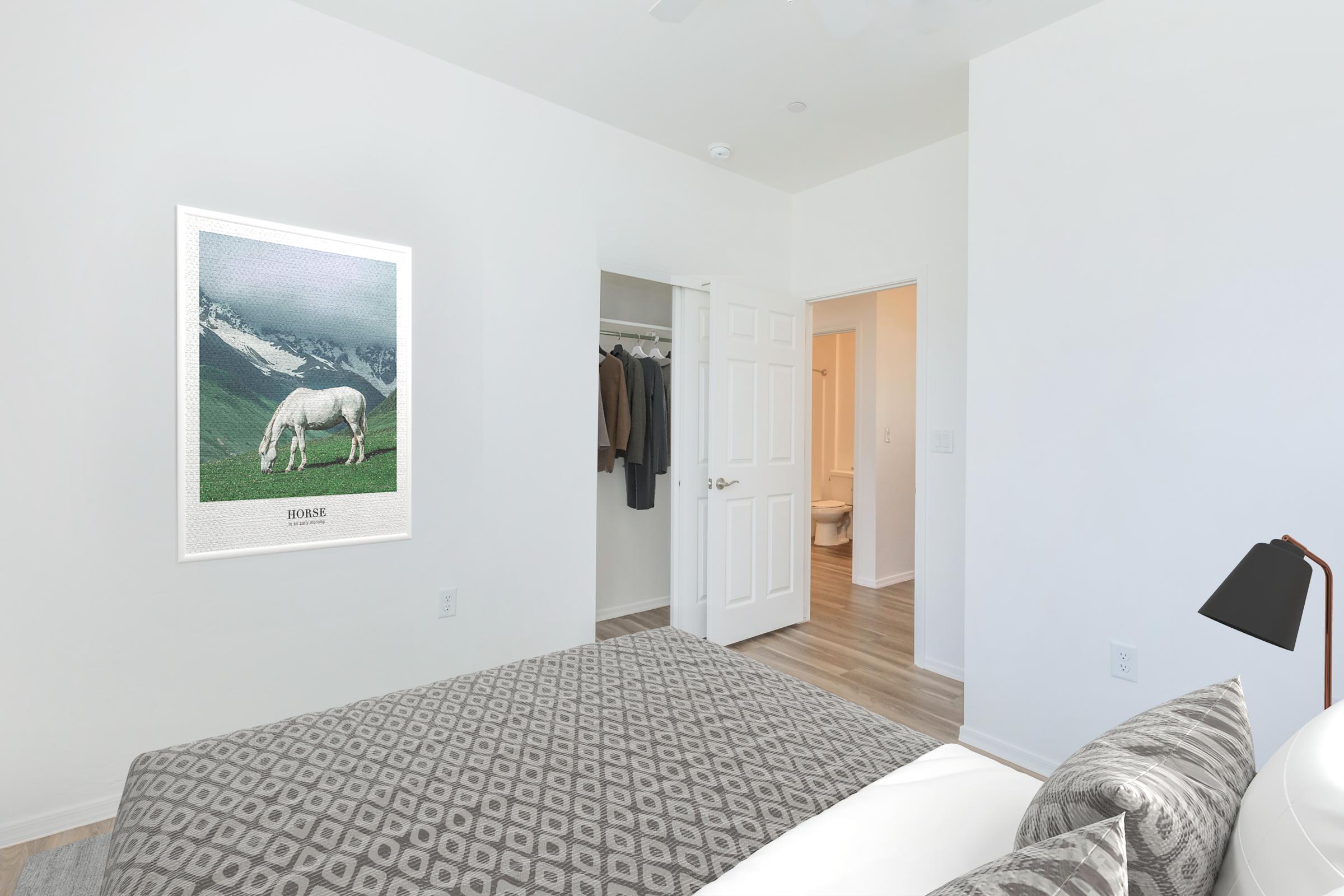 A minimalistic bedroom with a gray patterned bedspread. A framed poster of a horse in a green landscape hangs on the wall. A closet with clothes is visible in the background, and a bathroom doorway is partially seen. The room is bright and features light wooden flooring.