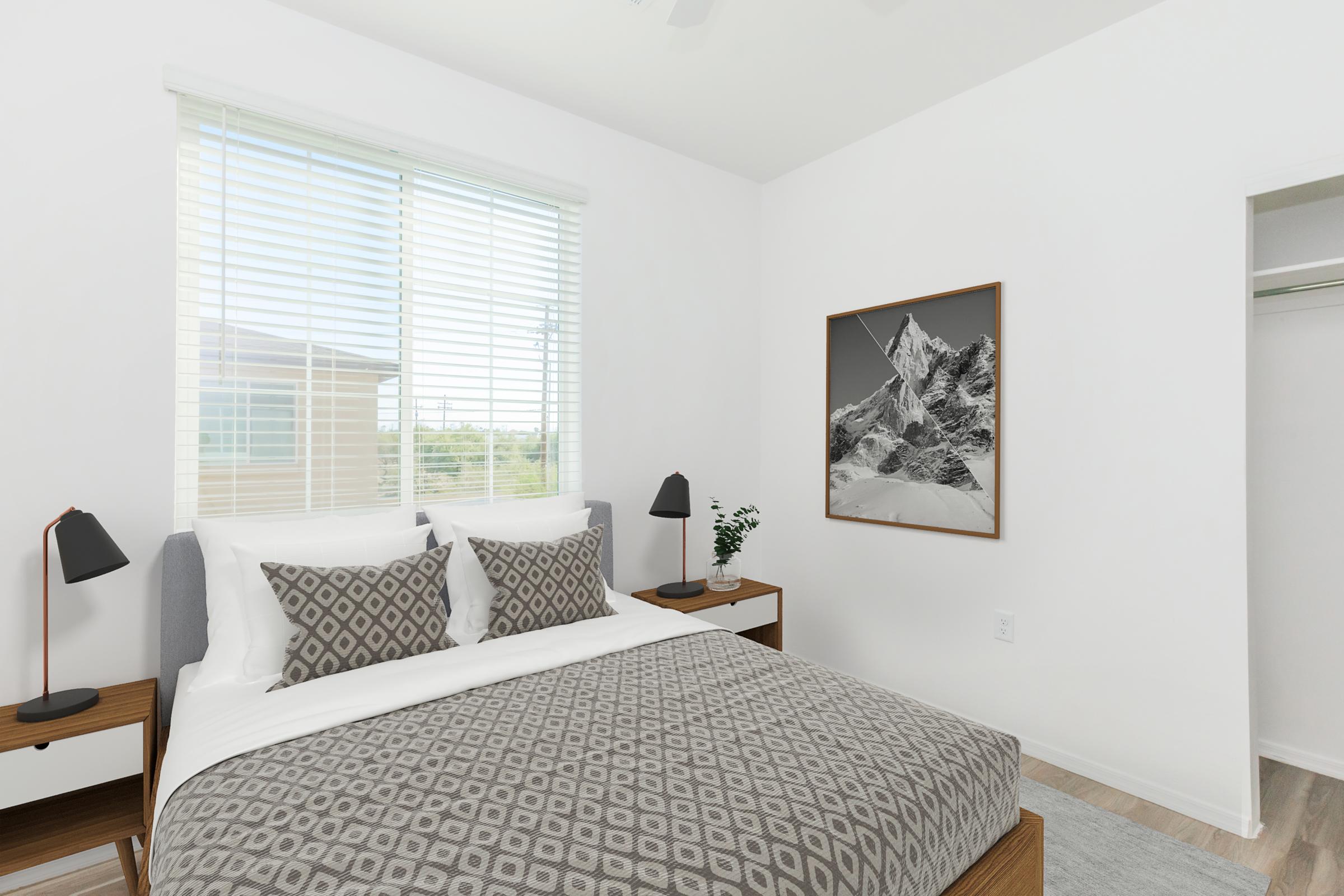 A modern bedroom featuring a neatly made bed with patterned bedding, accent pillows, and bedside tables with lamps. Natural light enters through a window with blinds. A framed black and white mountain photograph is hung on the wall, and a closet is partially visible in the corner.