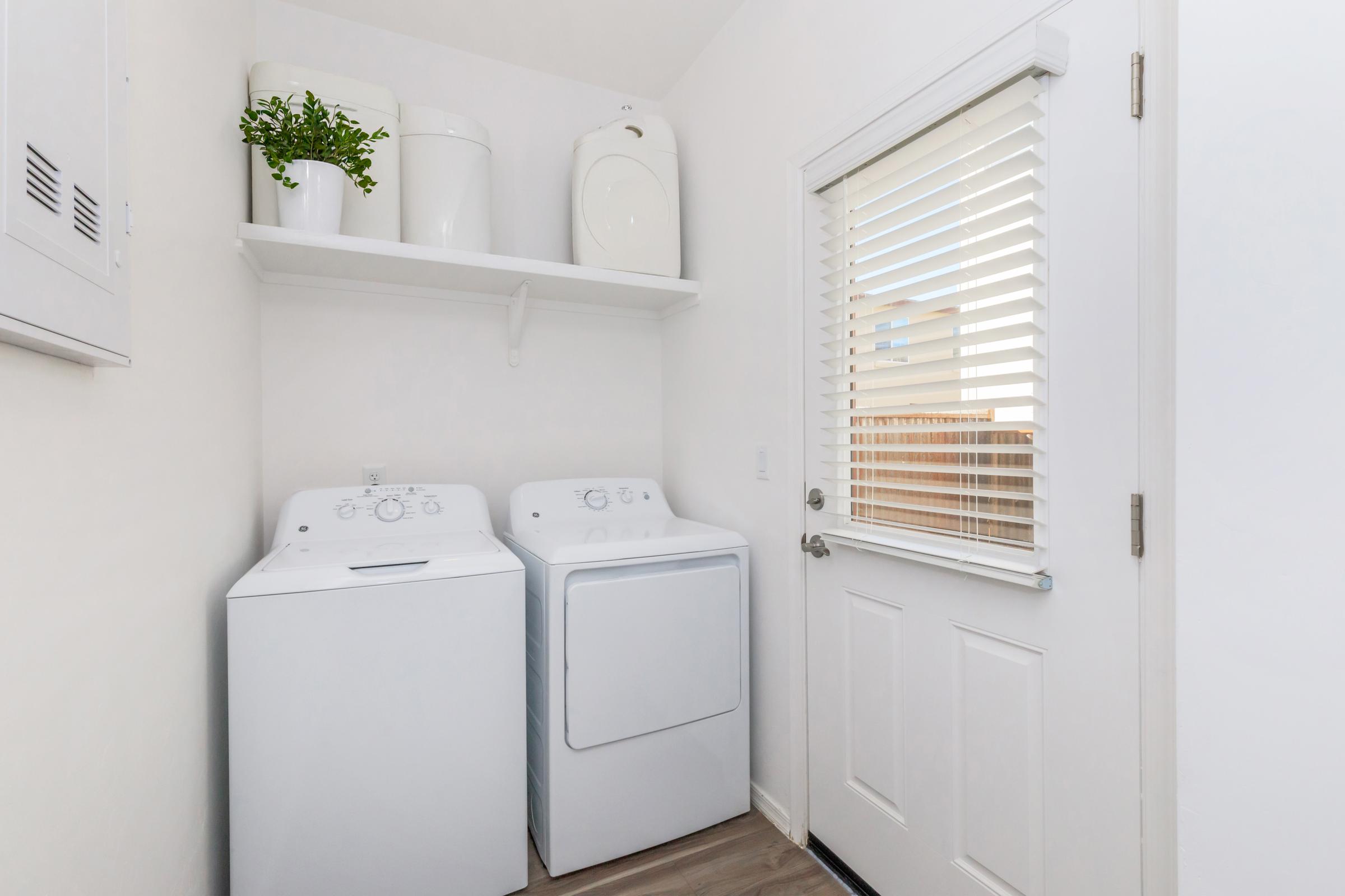 A bright laundry room featuring a white washing machine and dryer side by side, with a shelf above holding storage containers. A closed door with vertical blinds provides natural light, while the walls are painted white and the floor is wooden, creating a clean and organized space.