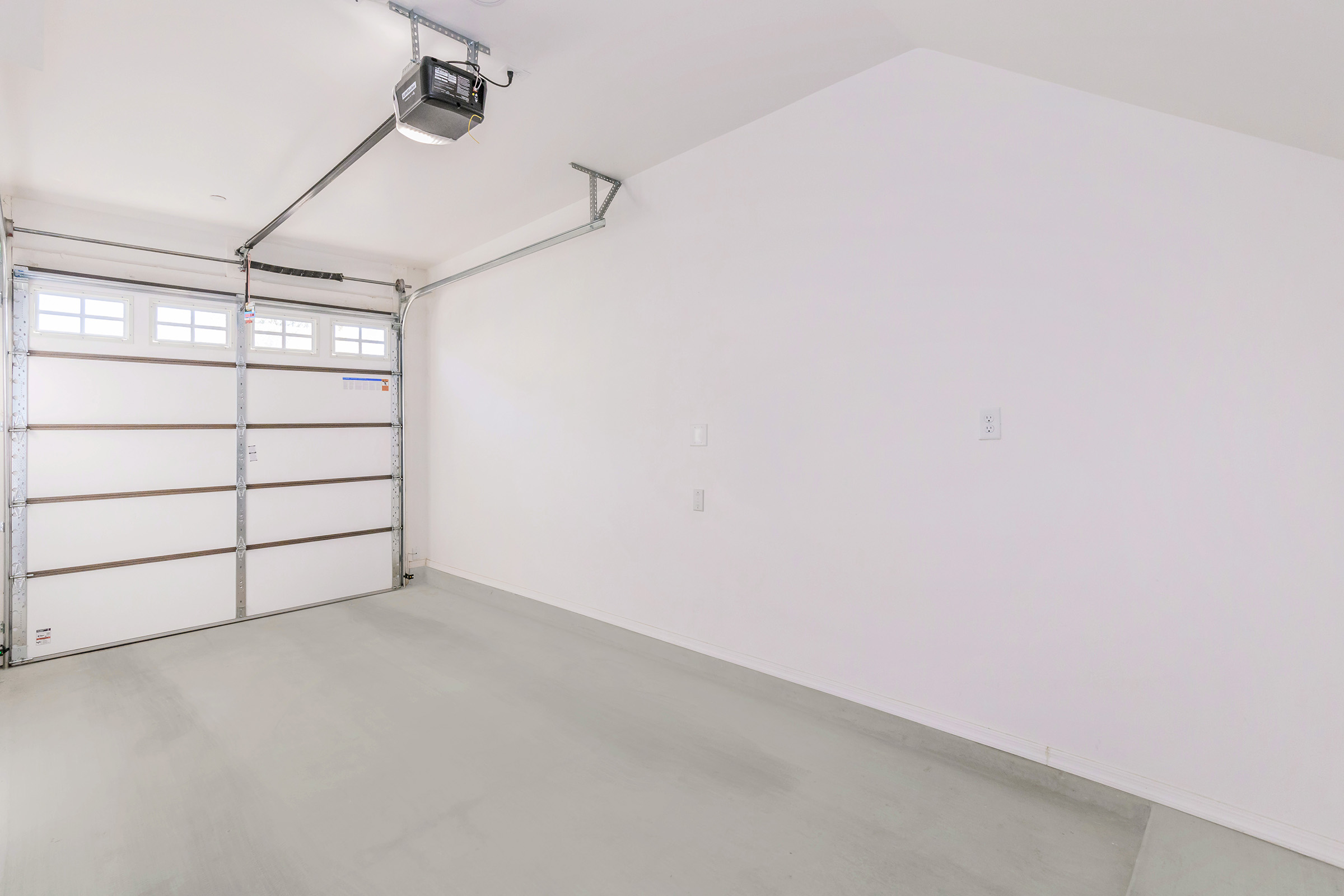 A clean and empty garage space featuring a large garage door on one side, white walls, and a smooth concrete floor. The garage is well-lit with natural light coming through the windows above the door, demonstrating a minimalist and uncluttered environment suitable for storage or parking.