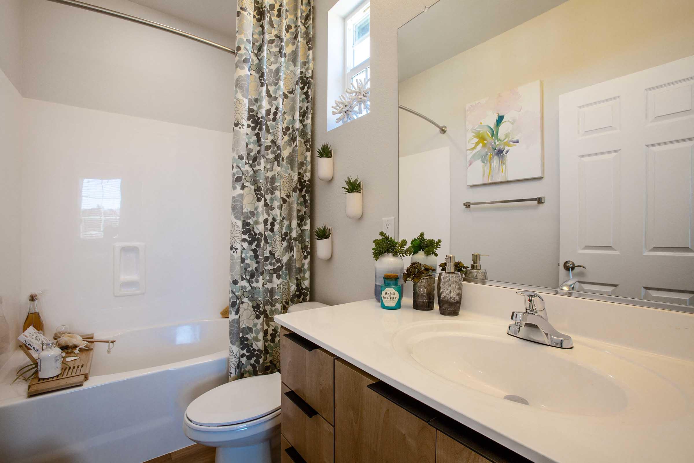 A modern bathroom featuring a bathtub with a shower curtain displaying a leafy pattern. A white sink sits on a wooden vanity, accompanied by decorative plants and toiletries. Natural light enters through a window, enhancing the bright ambiance, while a decorative painting decorates the wall above the sink.