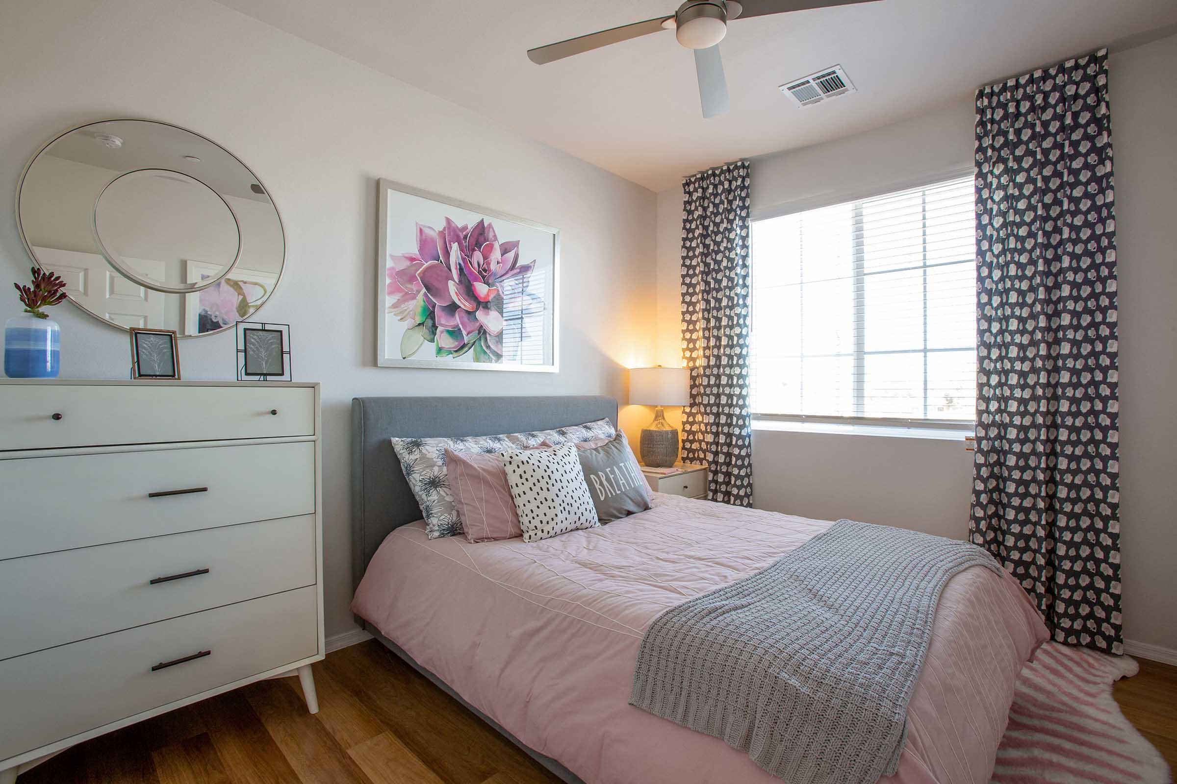 Cozy bedroom featuring a light pink bedspread and decorative pillows. There is a white dresser with framed pictures on top, a round mirror on the wall, and large windows with patterned curtains allowing natural light. A potted plant adds a touch of greenery to the room.