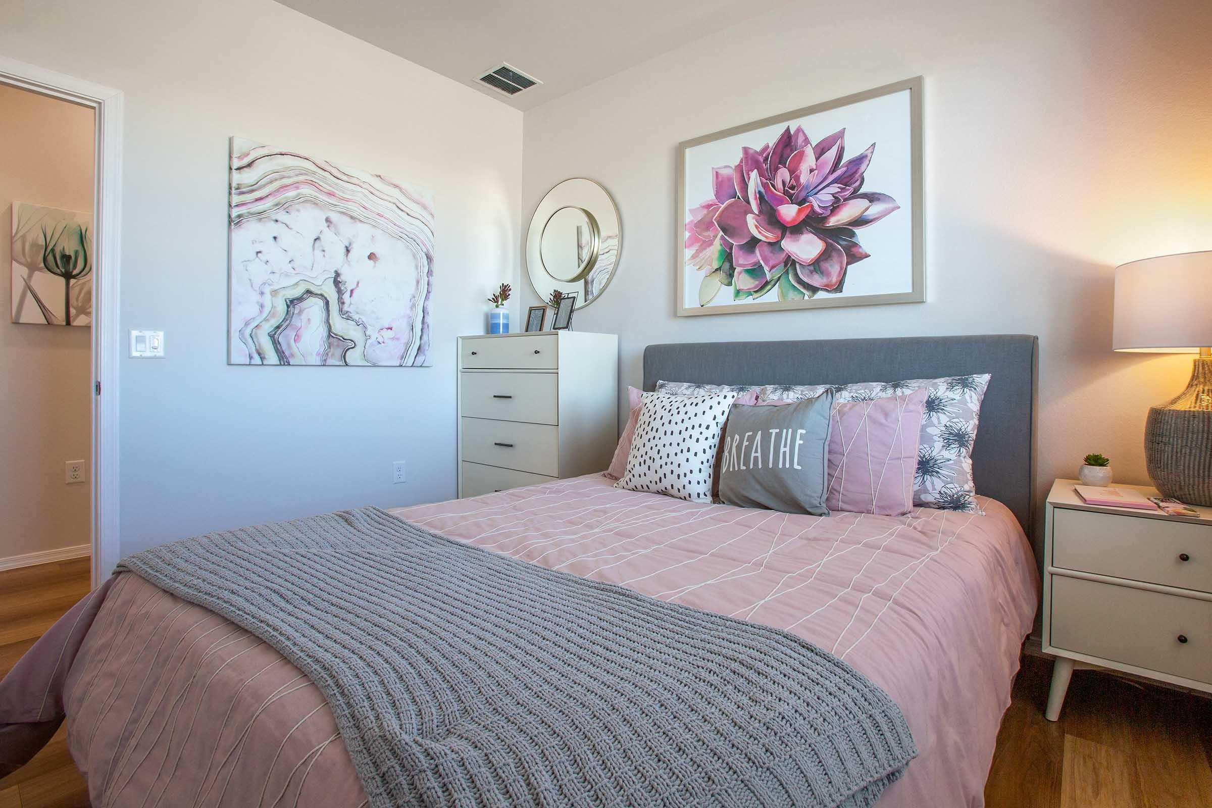 A cozy bedroom featuring a queen-sized bed with a light pink duvet and decorative pillows. The walls are adorned with two pieces of art: a floral painting and a marbled design. A modern nightstand with a lamp sits beside the bed, and there's a dresser on the opposite wall, creating a calm and inviting atmosphere.
