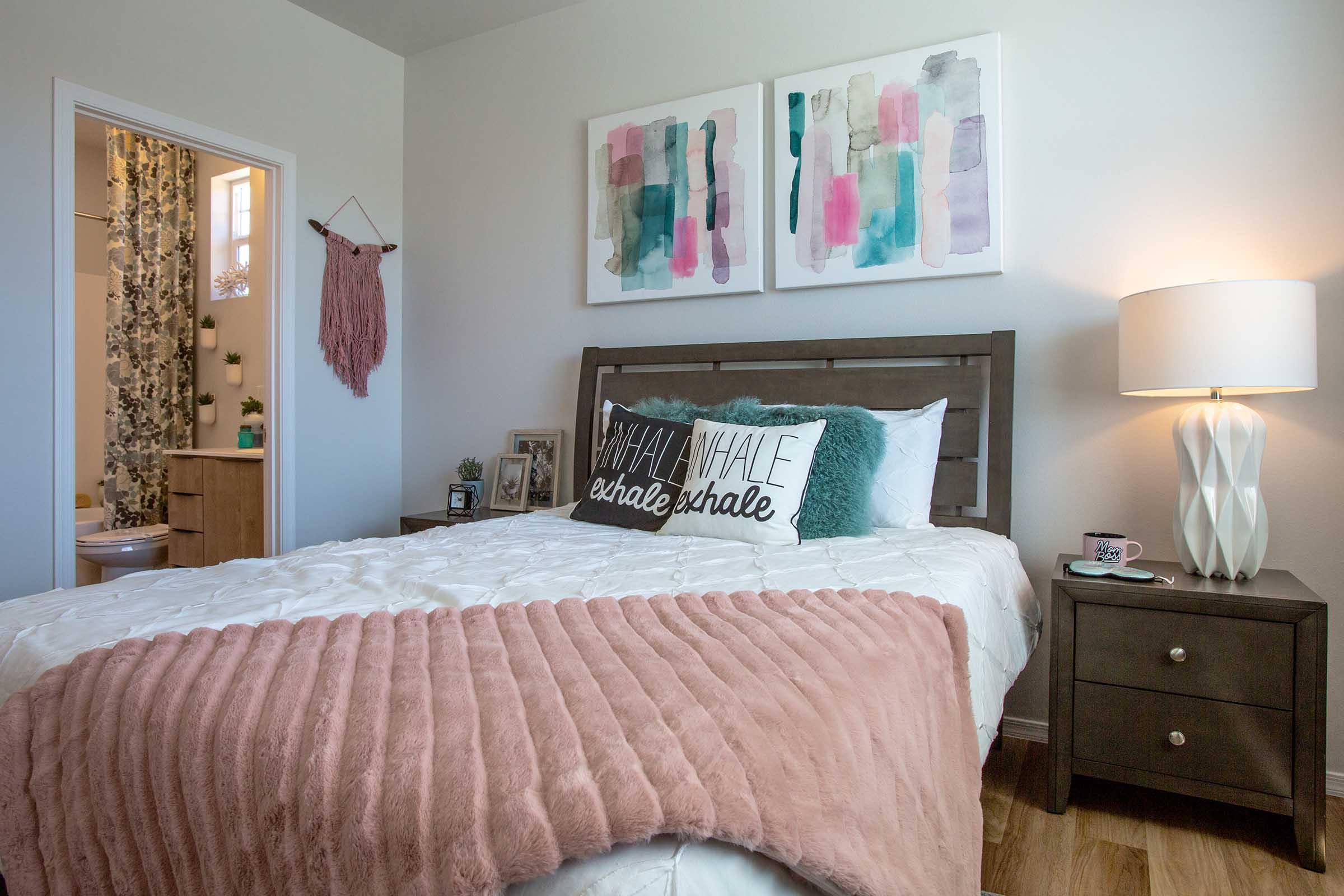 A cozy bedroom featuring a wooden bed with white bedding and a pink throw blanket. Two abstract paintings adorn the wall above the bed. A bedside table holds a lamp and decorative items. A bathroom door is visible in the background, along with a wall hanging and green plants.