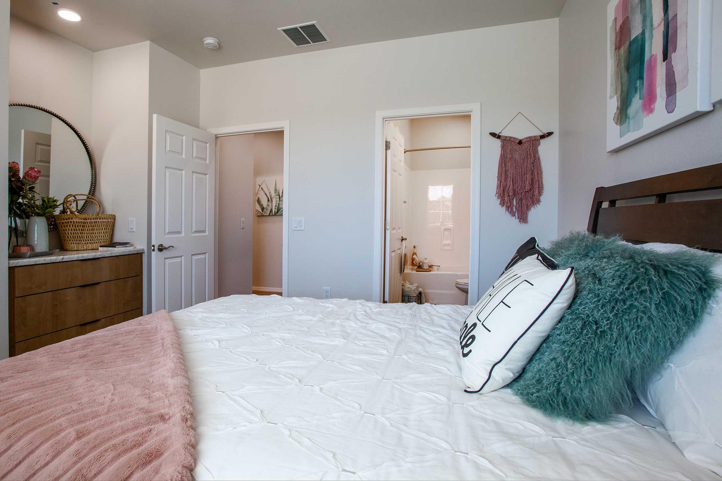 A cozy bedroom featuring a bed with a fluffy teal throw pillow and patterned black and white pillows. There's a wooden dresser with a mirror, a plant, and a decorative basket. Two doorways lead to a hallway and a bathroom. Soft lighting creates a warm, inviting atmosphere.