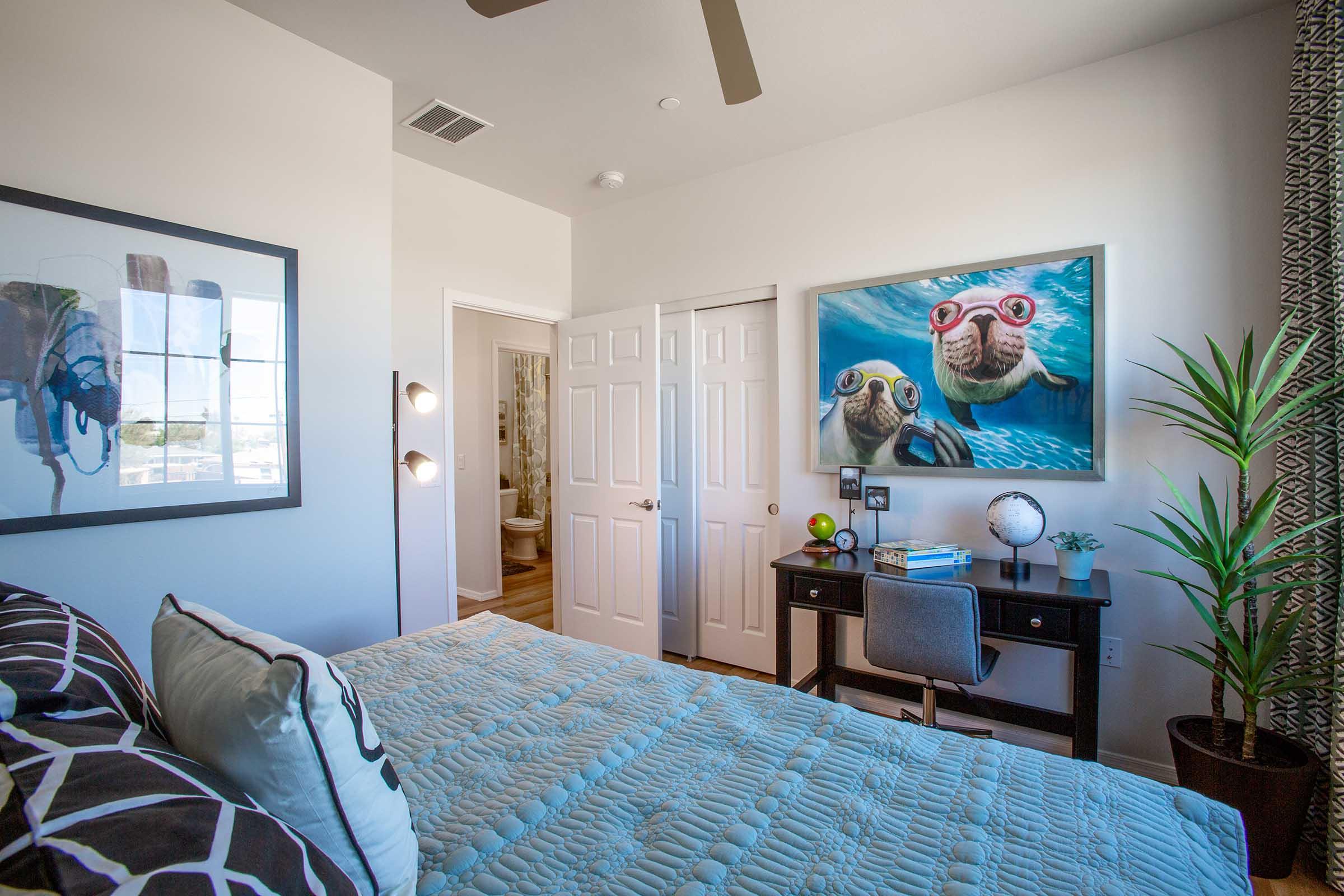 A modern bedroom featuring a queen-sized bed with a blue quilt, a stylish desk with a chair, and a large wall art of two playful sea otters wearing goggles. The room has light-colored walls, a plant in the corner, and a door leading to a bathroom in the background. A ceiling fan adds to the ambiance.