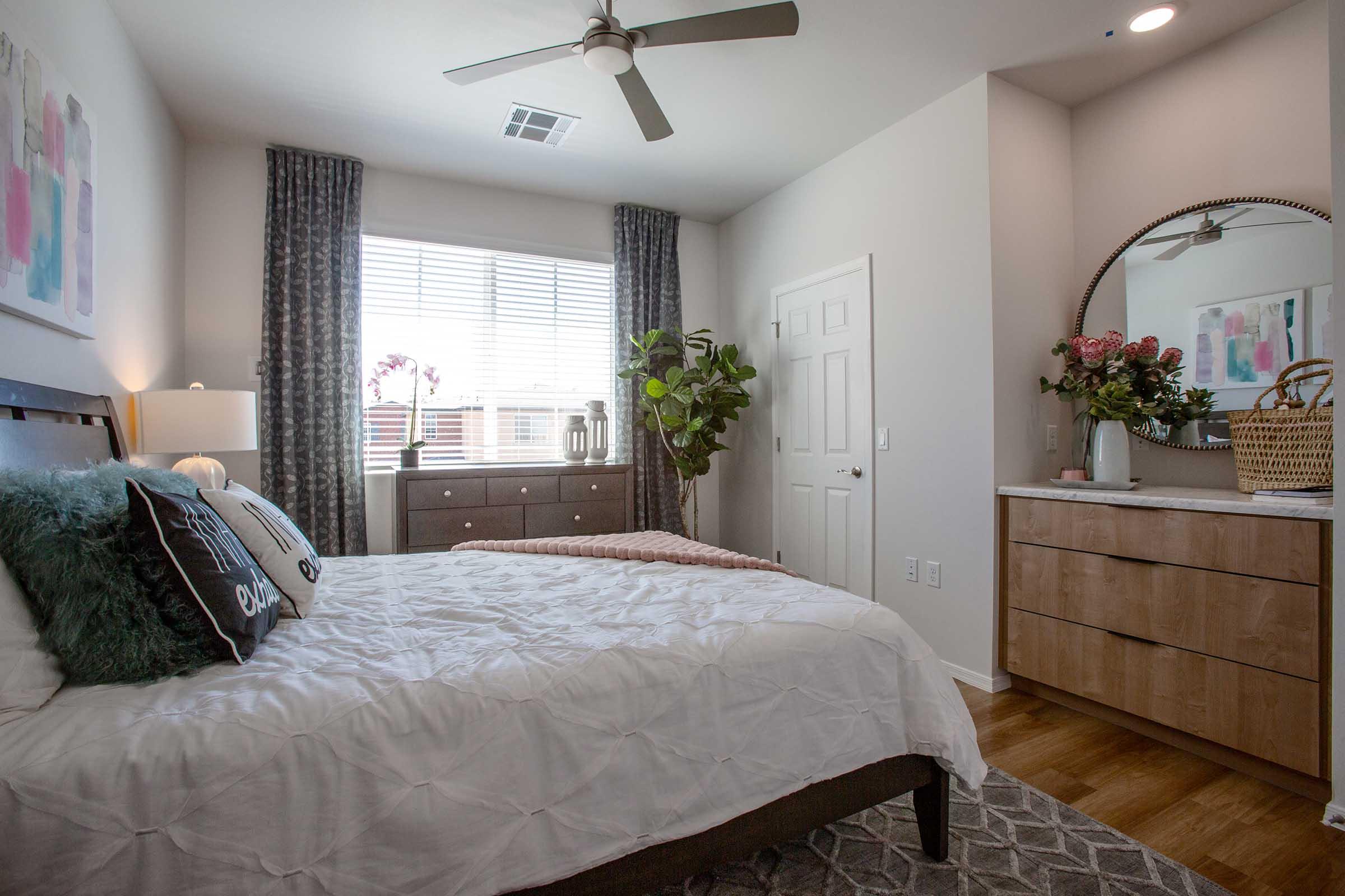 A cozy bedroom featuring a queen-sized bed with a white quilt and decorative pillows. Natural light streams in through a window with sheer curtains, illuminating a plant beside the bed. A wooden dresser and a wicker basket with flowers are also visible, creating a warm and inviting atmosphere.
