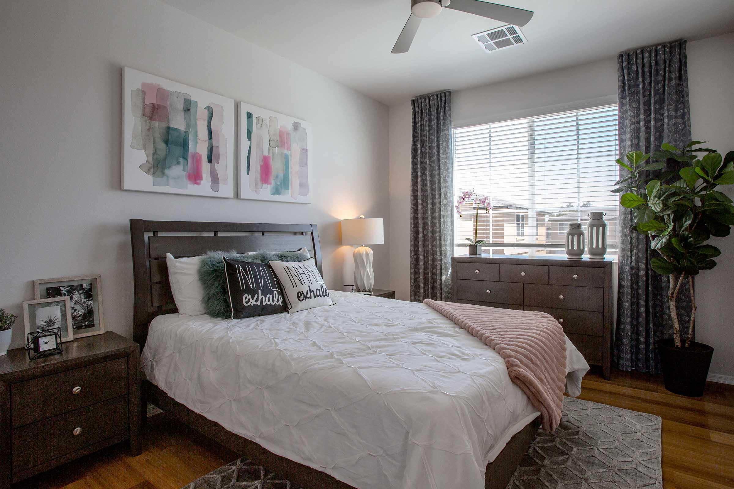 A well-decorated bedroom featuring a wooden bed frame with white bedding and accent pillows. There's a stylish nightstand, a lamp, and a dresser with decor items. Large windows allow natural light, and a potted plant adds a touch of greenery. Abstract artwork in pastel colors hangs on the wall.