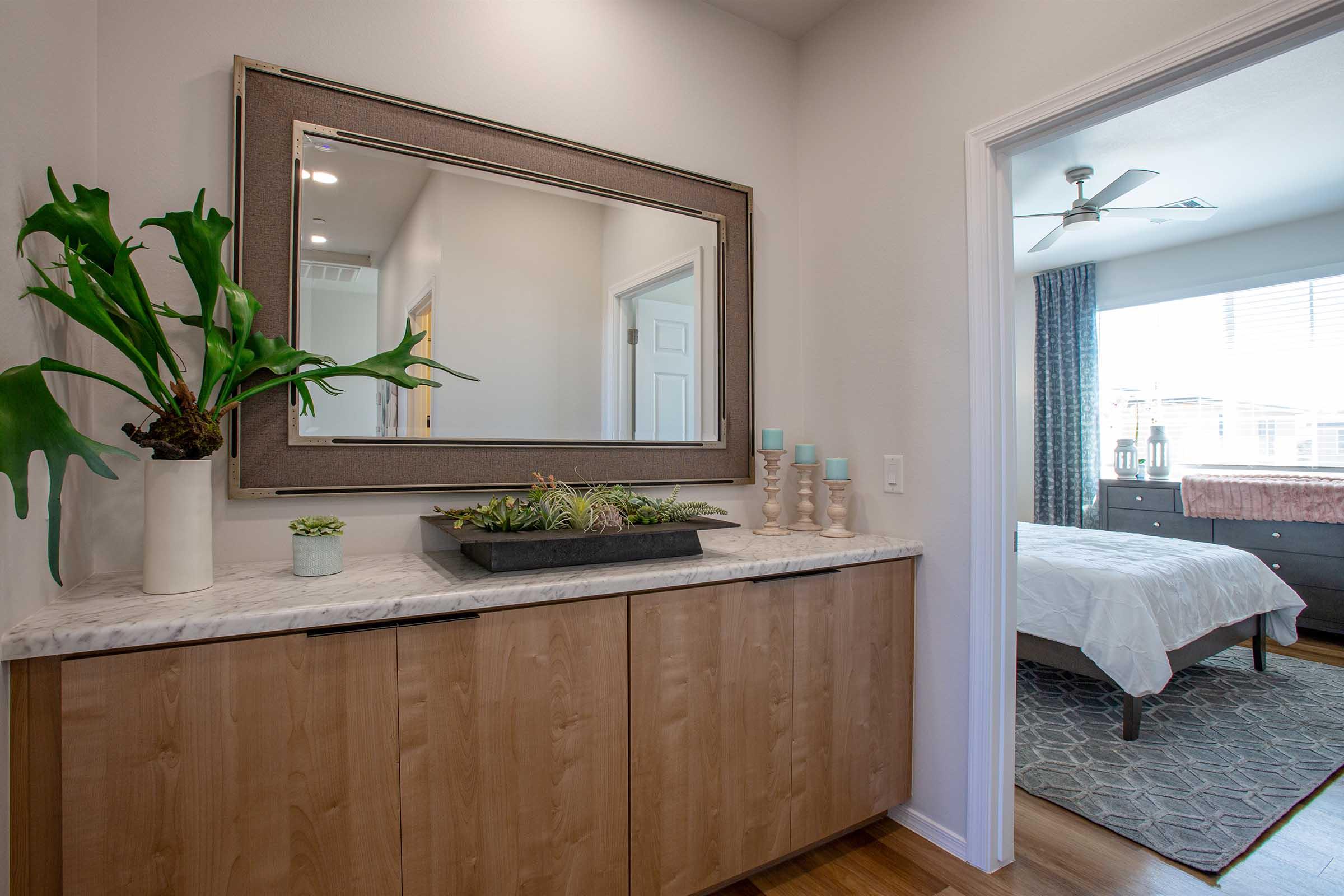 A modern interior space featuring a stylish sideboard with a marble top, adorned with a decorative planter and candles. A large mirror hangs above the sideboard, reflecting the light. In the background, a bedroom is visible, complete with a bed and patterned rug, enhancing the inviting atmosphere.