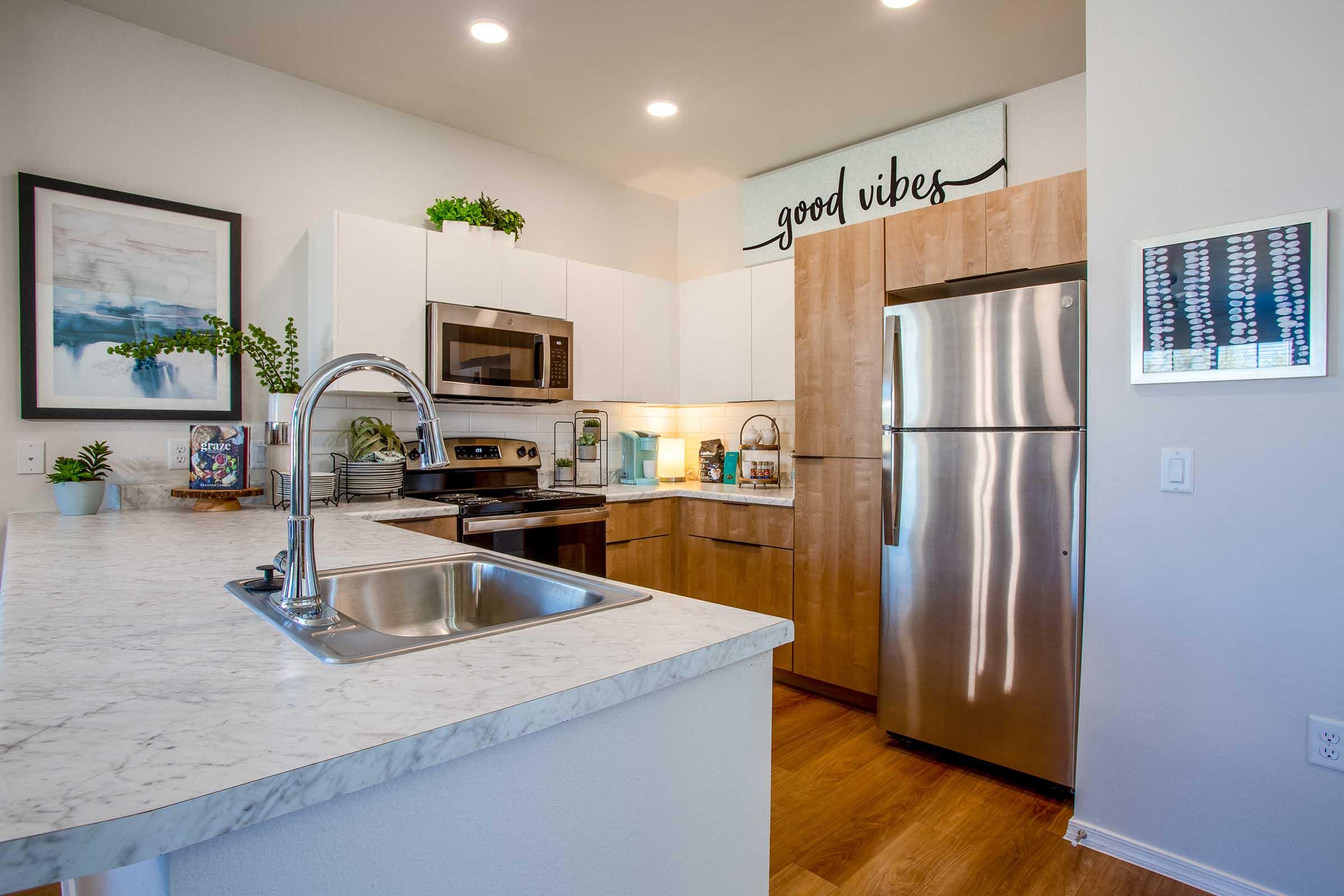 Modern kitchen featuring a marble countertop, stainless steel appliances including a microwave and refrigerator, and wooden cabinetry. The wall displays the phrase "good vibes" above the sink. Decorative plants and artwork add a fresh touch to the space, which has warm wooden flooring and bright, welcoming lighting.