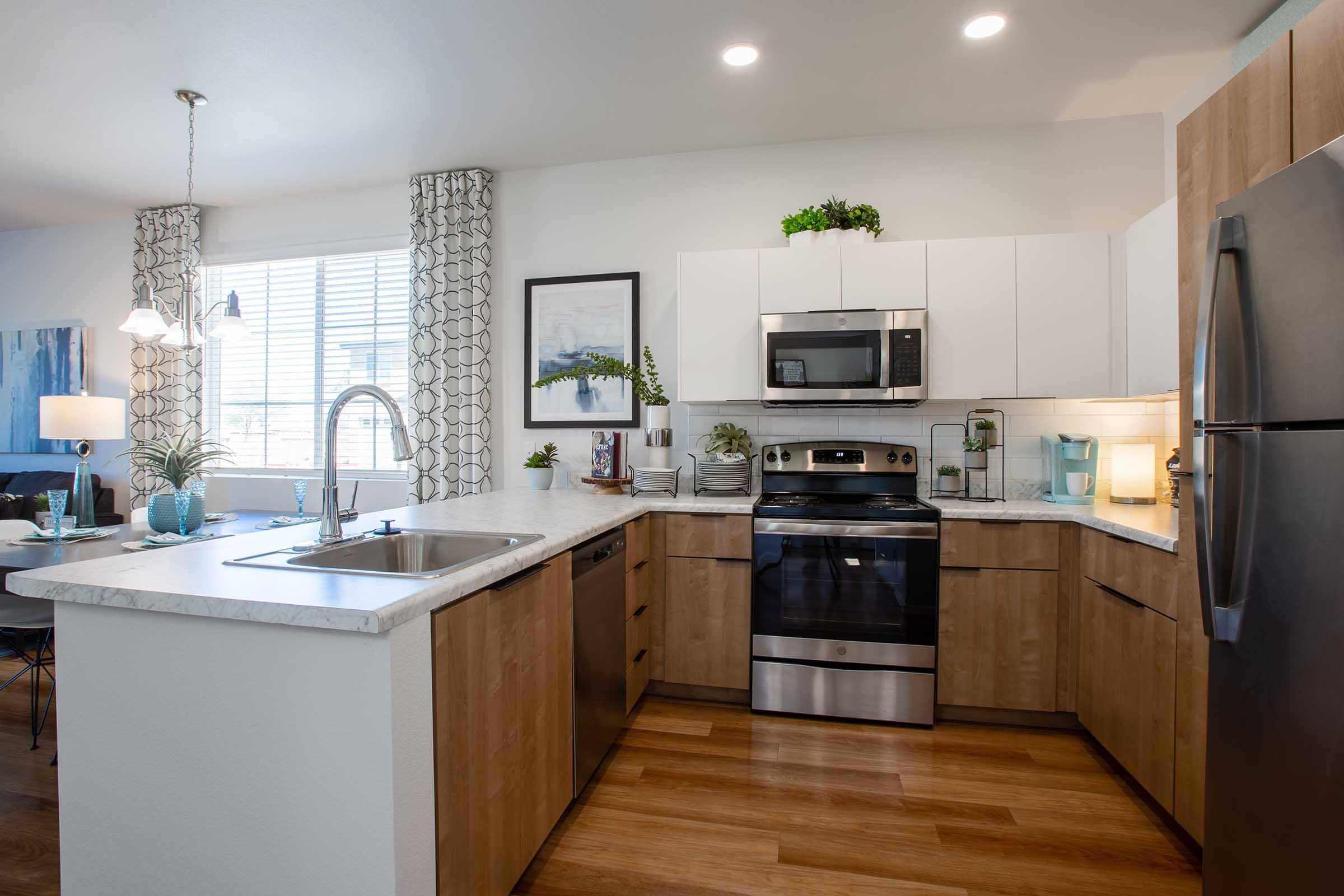 A modern kitchen featuring light wood cabinetry, a stainless steel stove, and a microwave. The countertop is made of white marble, and there's a double sink with a sleek faucet. Natural light floods the space through a window adorned with curtains. Decorative plants and kitchen essentials are placed around the room, creating a bright and inviting atmosphere.