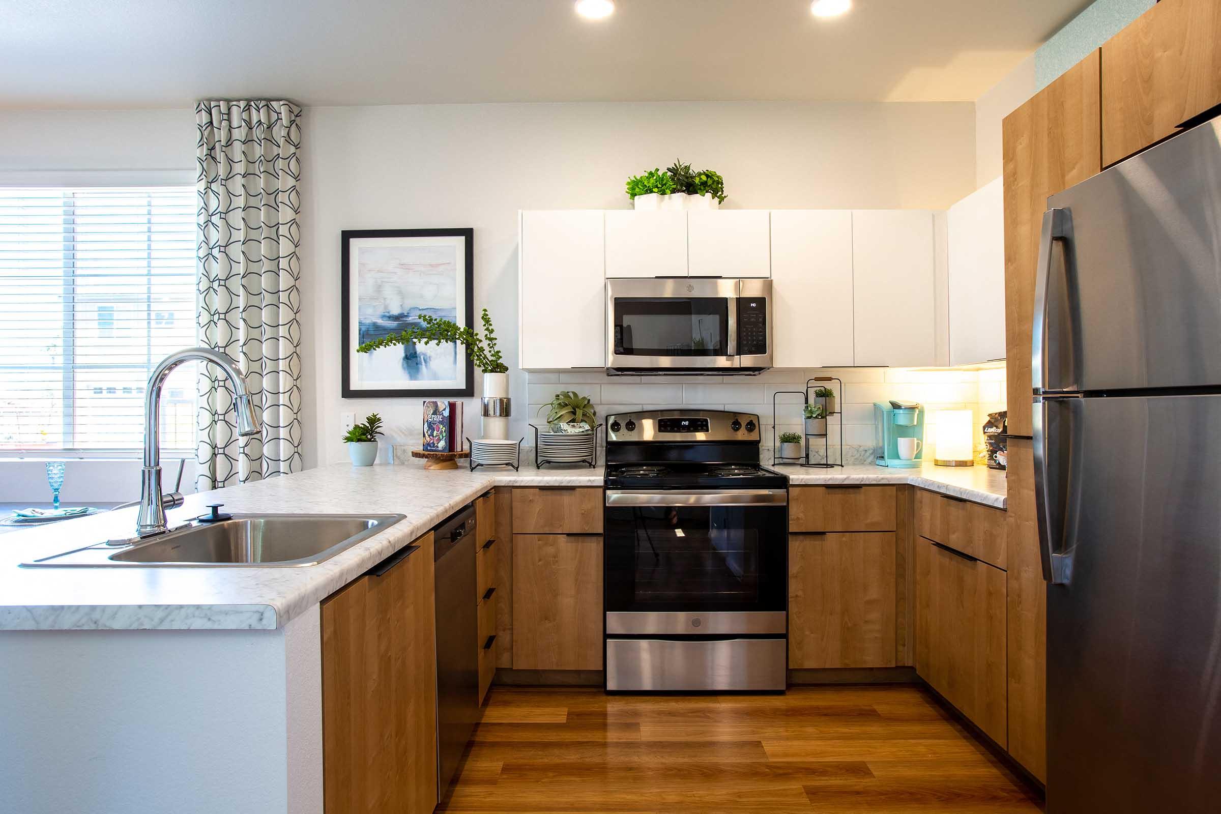 Modern kitchen featuring wooden cabinets, a stainless steel refrigerator and oven, a microwave, and a sink. The countertop is marble with plants and kitchen accessories. A large window provides natural light, complemented by a decorative curtain and a piece of wall art.
