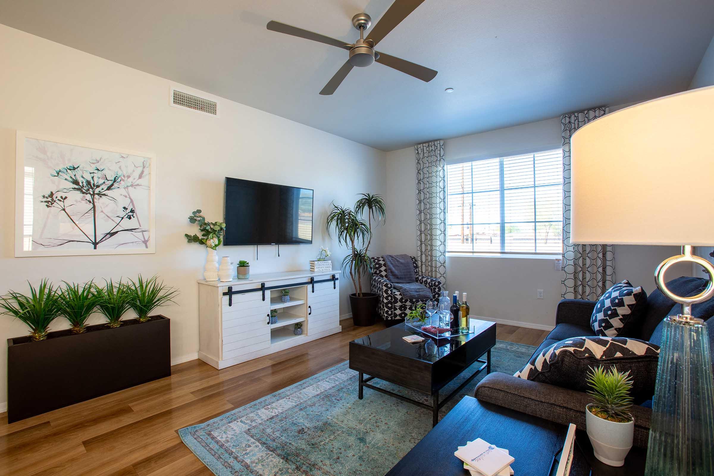 A modern living room featuring a comfortable gray sofa, a coffee table with a decorative centerpiece, a wall-mounted TV, and a stylish floor lamp. Large windows let in natural light, and potted plants add a touch of greenery. A patterned rug complements the wooden flooring.