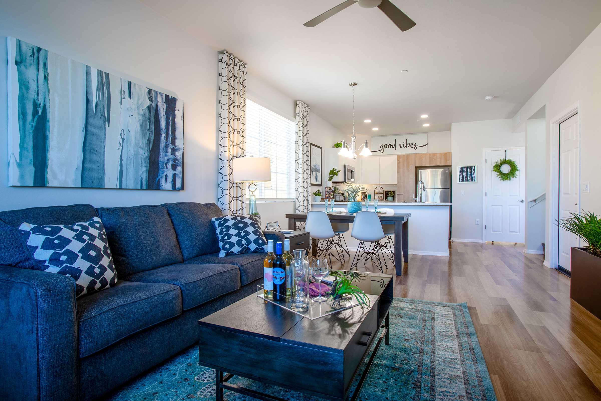A modern living room featuring a dark blue couch with patterned pillows, a coffee table with a decorative tray, and a dining area with white chairs. The space is brightened by large windows and stylish decor, including artwork on the walls and plants, creating a welcoming atmosphere.