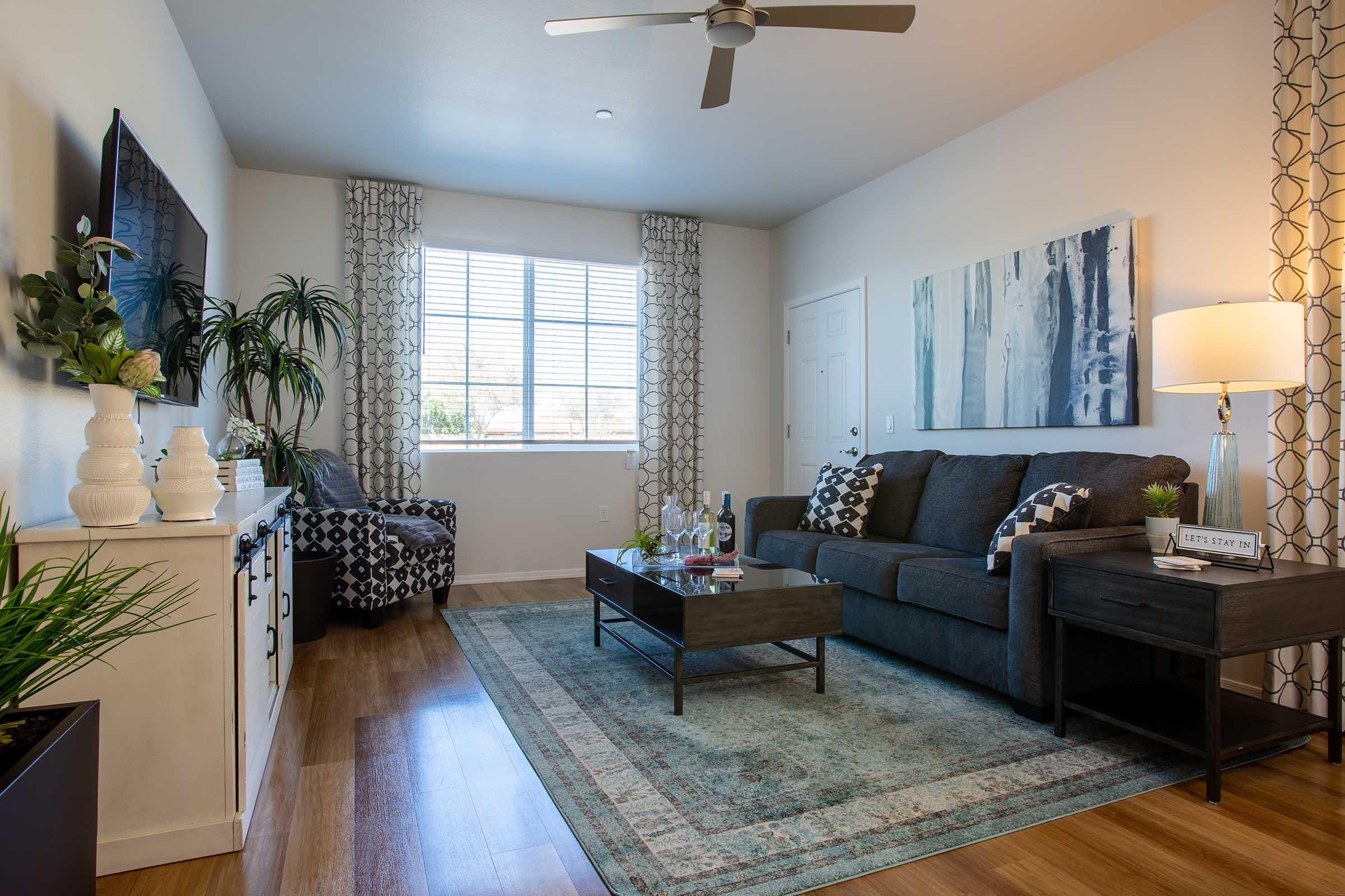A modern living room featuring a dark gray sofa, a coffee table with decorative items, and a patterned area rug. Soft lighting from a lamp on a nearby table accentuates the space, which includes a TV on the wall and large windows with sheer curtains, allowing natural light to fill the room.