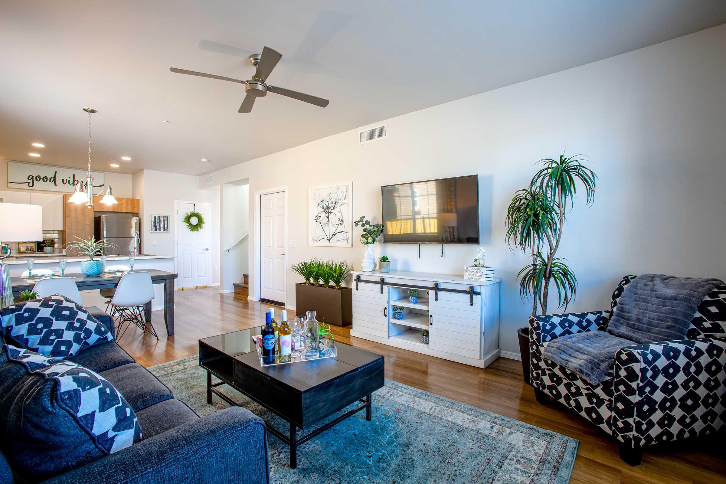 A bright and inviting living room featuring a cozy sofa and armchair, a coffee table with a selection of drinks, and a wall-mounted television. The space includes a kitchen area in the background with modern appliances, decorative plants, and wall art, creating a warm and welcoming atmosphere.
