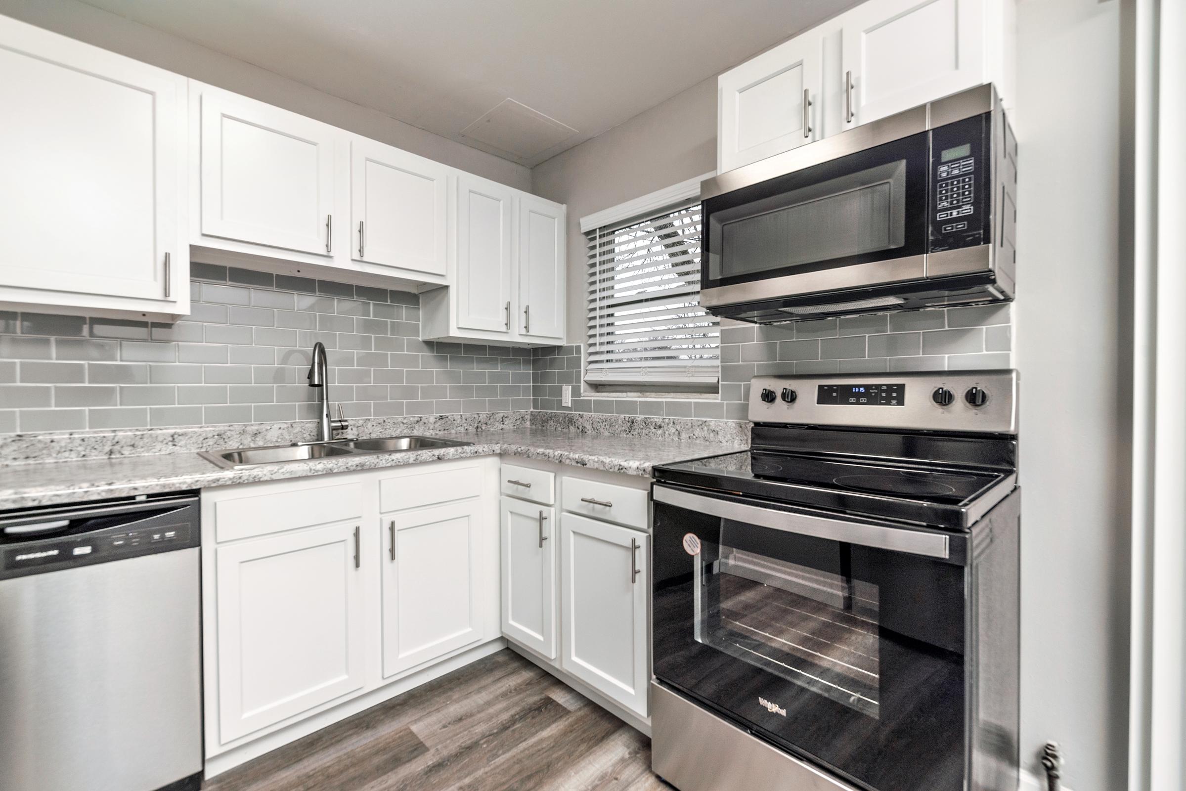 Modern kitchen featuring white cabinetry, a stainless steel microwave, and a black oven. Gray subway tile backsplash complements the granite countertops. The space includes a stainless steel dishwasher and a sleek stainless sink, with natural light coming in from a window.