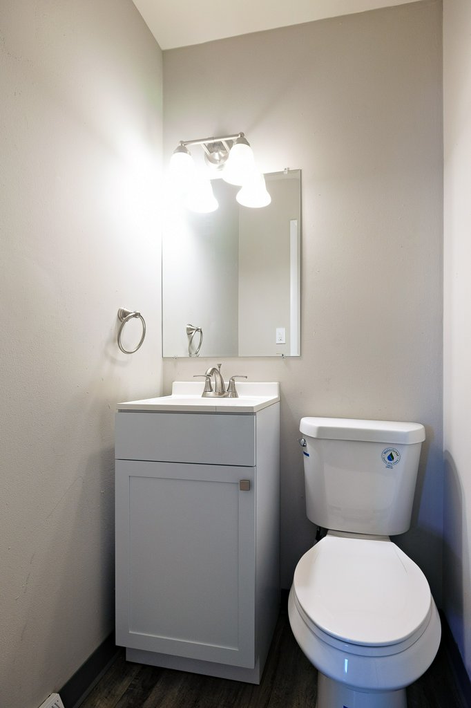 A small bathroom featuring a white vanity with a sink, a wall-mounted mirror above it, a toilet to the right, and a wall light fixture with three bulbs. The walls are painted a light gray, and the flooring is a dark, wood-like material.