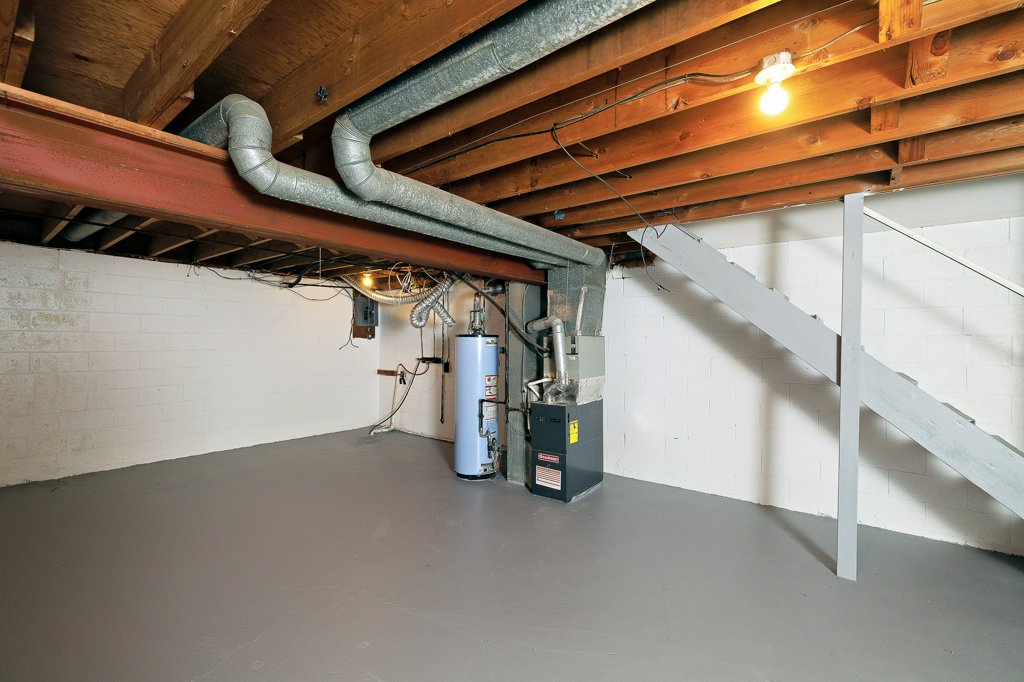 A clean, unfinished basement featuring exposed wooden beams and concrete walls. Along one wall, there are pipes and ductwork. A blue water heater and a black furnace are positioned beside each other. A set of stairs leads to an upper level, and a single light bulb illuminates the space. The floor is painted grey.