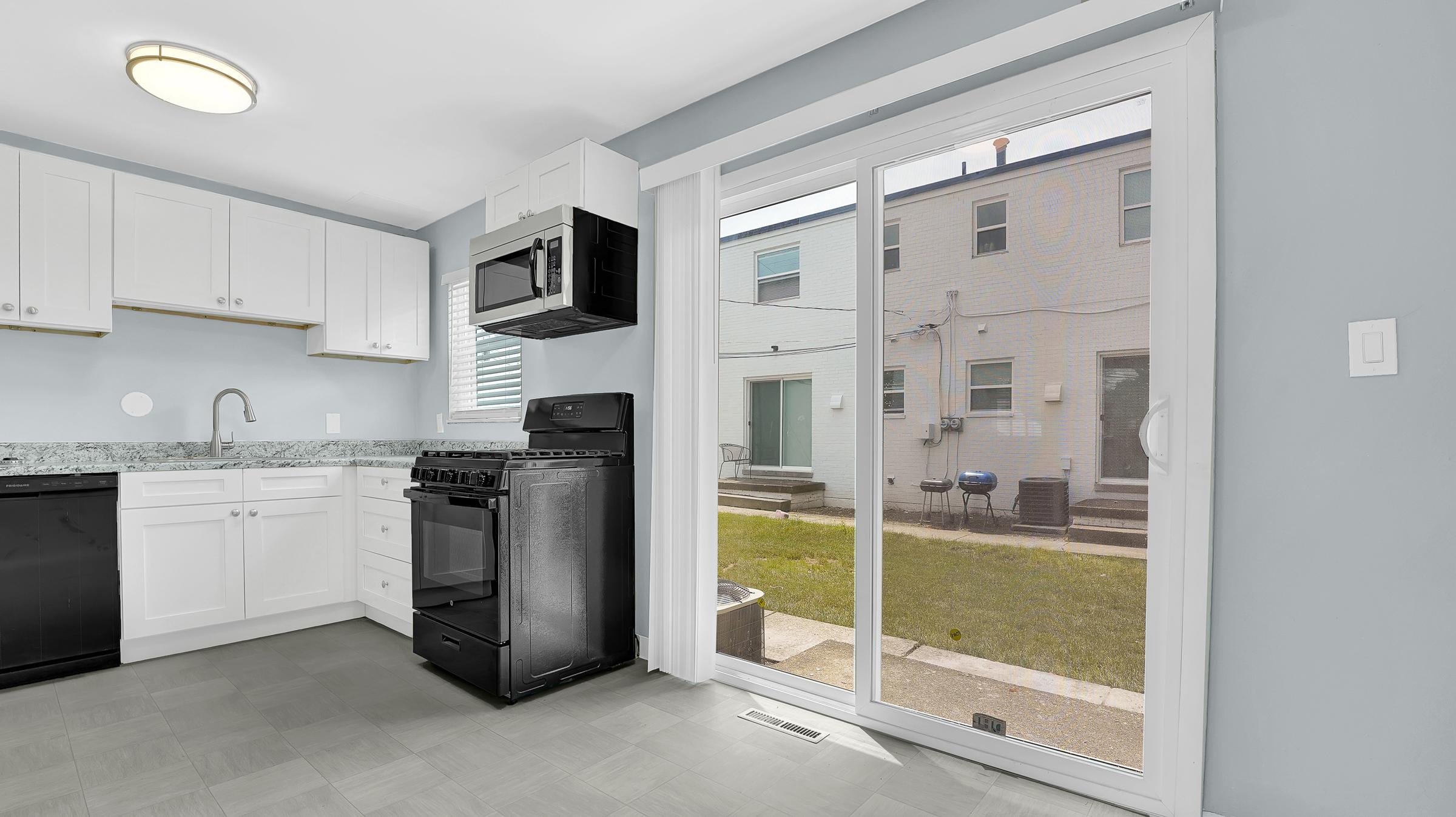 A modern kitchen featuring white cabinetry, a black stove and microwave, and a sink. A sliding glass door opens to an outdoor area with grass and stairs leading to another level. Natural light illuminates the space through a large window. The floor is light gray tile.