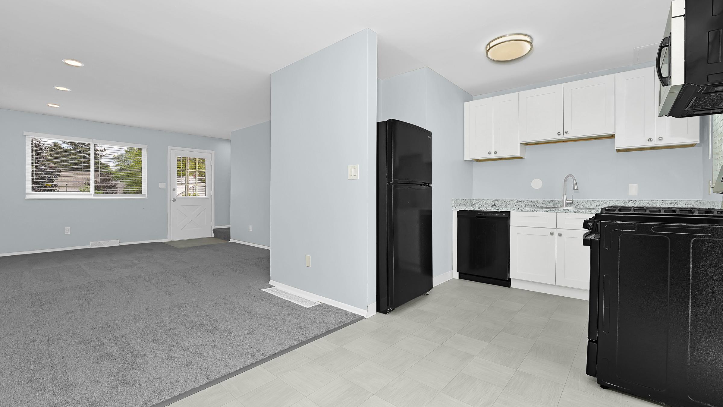A modern kitchen and living area featuring light blue walls, white cabinets, and stainless steel appliances. The kitchen includes a black refrigerator, a black stove, and granite countertops. The adjacent living space has gray carpeting and a window allowing natural light. A door leads outside.
