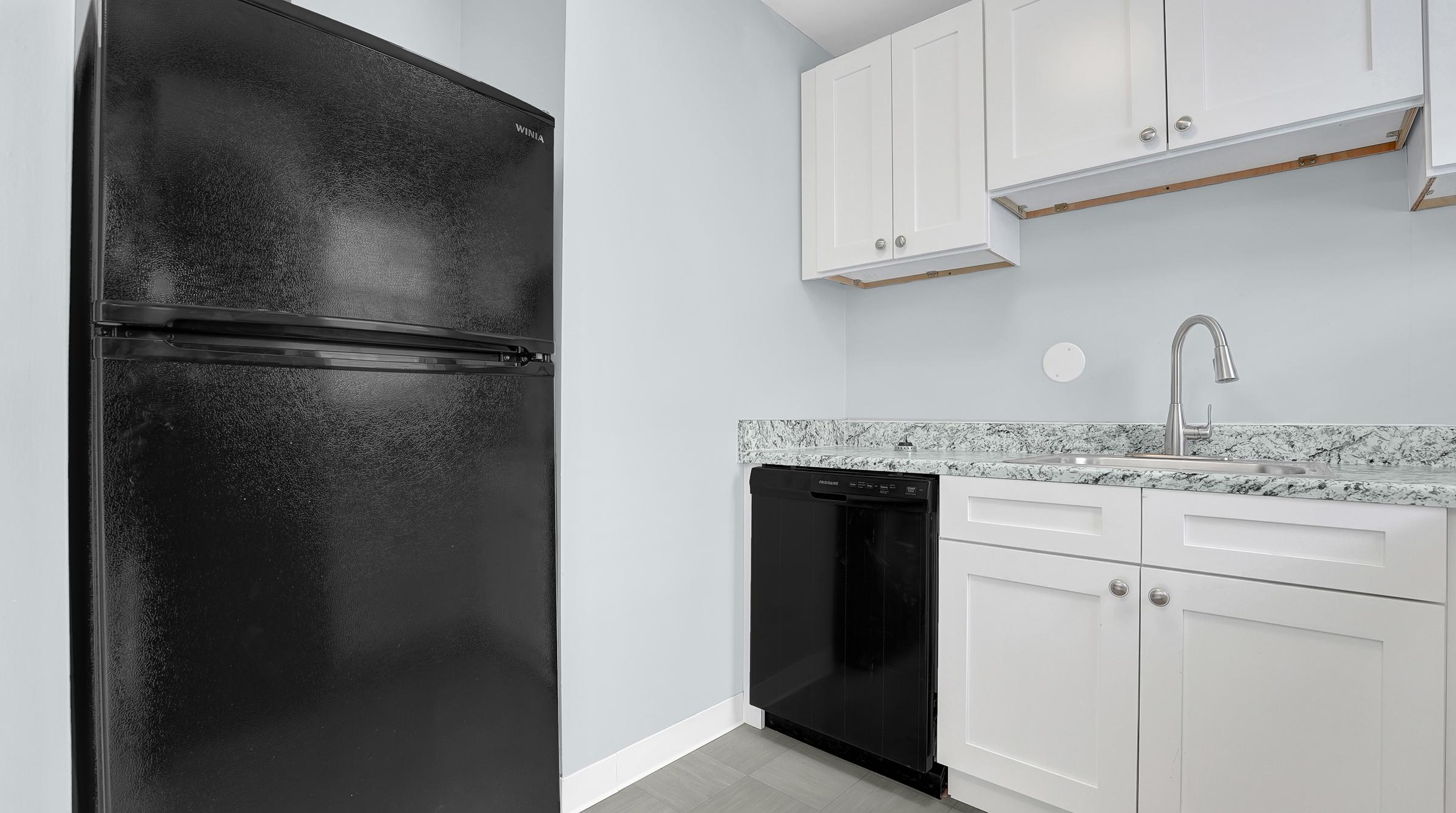 A kitchen area featuring a black refrigerator and a black dishwasher integrated into white cabinetry. The countertop is a speckled design in shades of grey and white, with a stainless steel faucet installed above the sink. The walls are painted light blue, creating a modern and clean aesthetic.