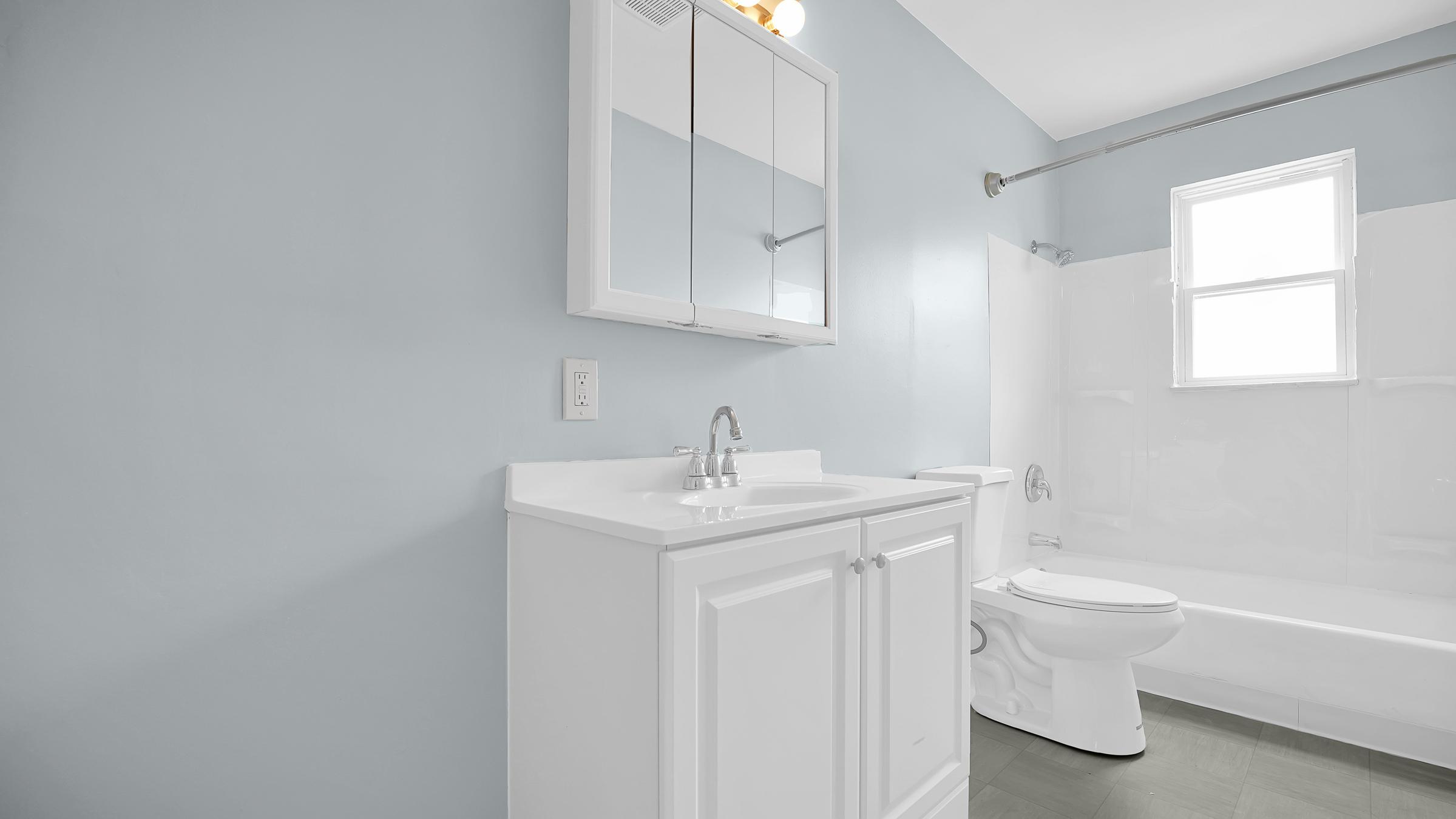 A modern bathroom featuring mint green walls, a white vanity with two cabinets, a mirror above the sink, a bathtub with a shower, and a toilet. Natural light enters through a window, highlighting the clean and minimalist design.