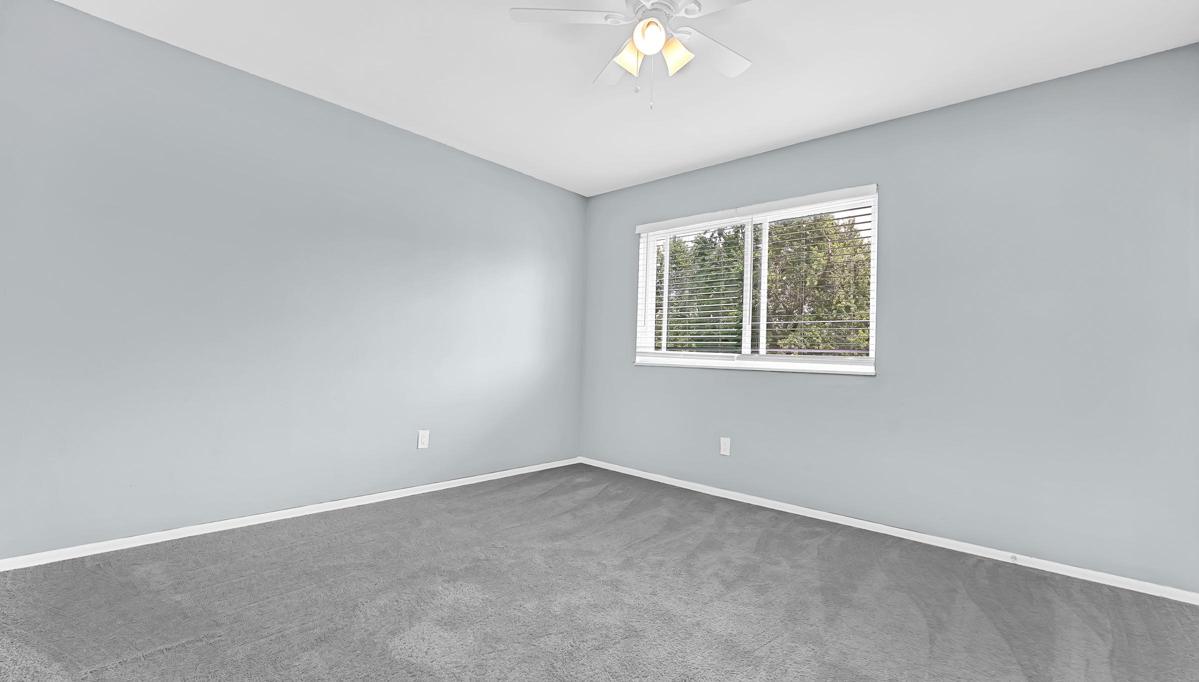 A small, sunny room with light blue walls and a window dressed with white blinds. The floor is covered with gray carpet, and a ceiling fan is visible. The space appears empty and clean, ideal for personal customization or furnishing.