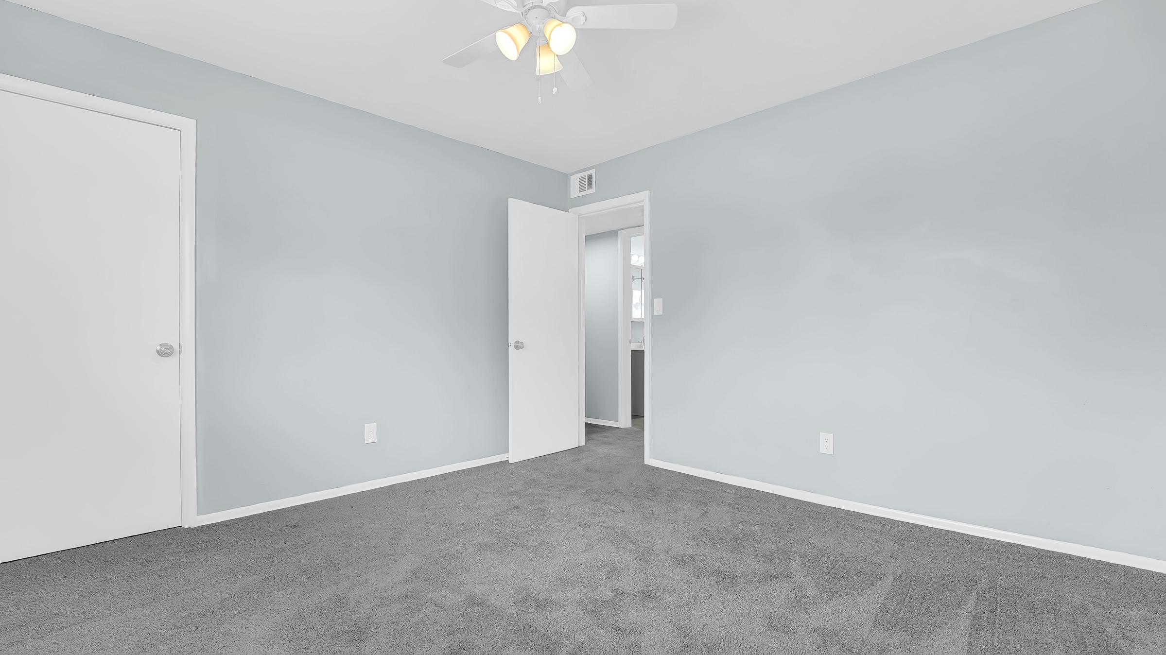 A view of a small, empty room with light blue walls and gray carpet. There's a white ceiling fan with four lights. A closed white door is on the left, and an open door leading to another room is on the right, revealing a glimpse of the adjacent space.