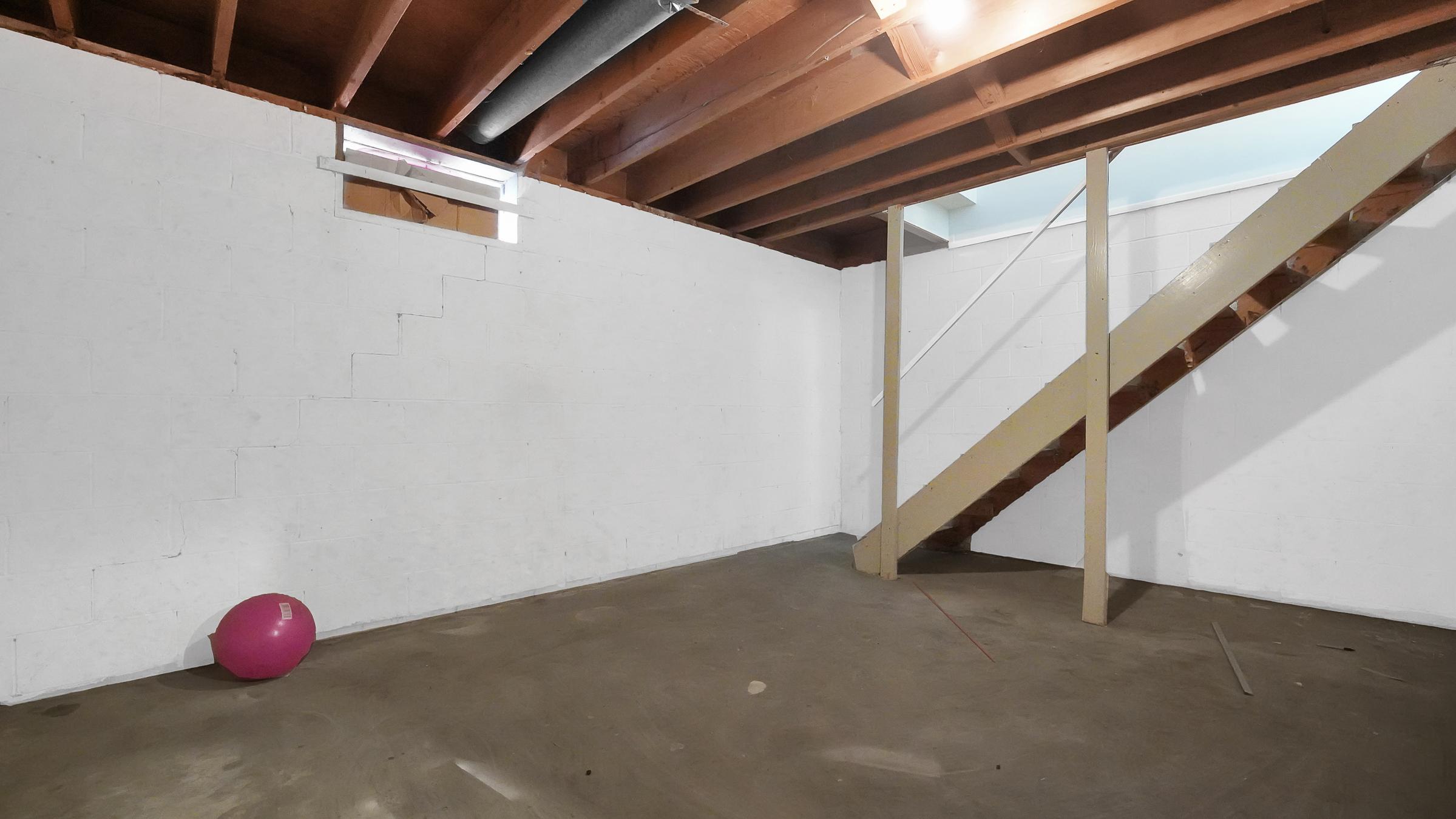 Empty basement with white walls and exposed wooden beams. A staircase in the corner leads up, and a pink exercise ball is resting on the concrete floor. The room is well-lit from overhead fixtures, creating a spacious feel.