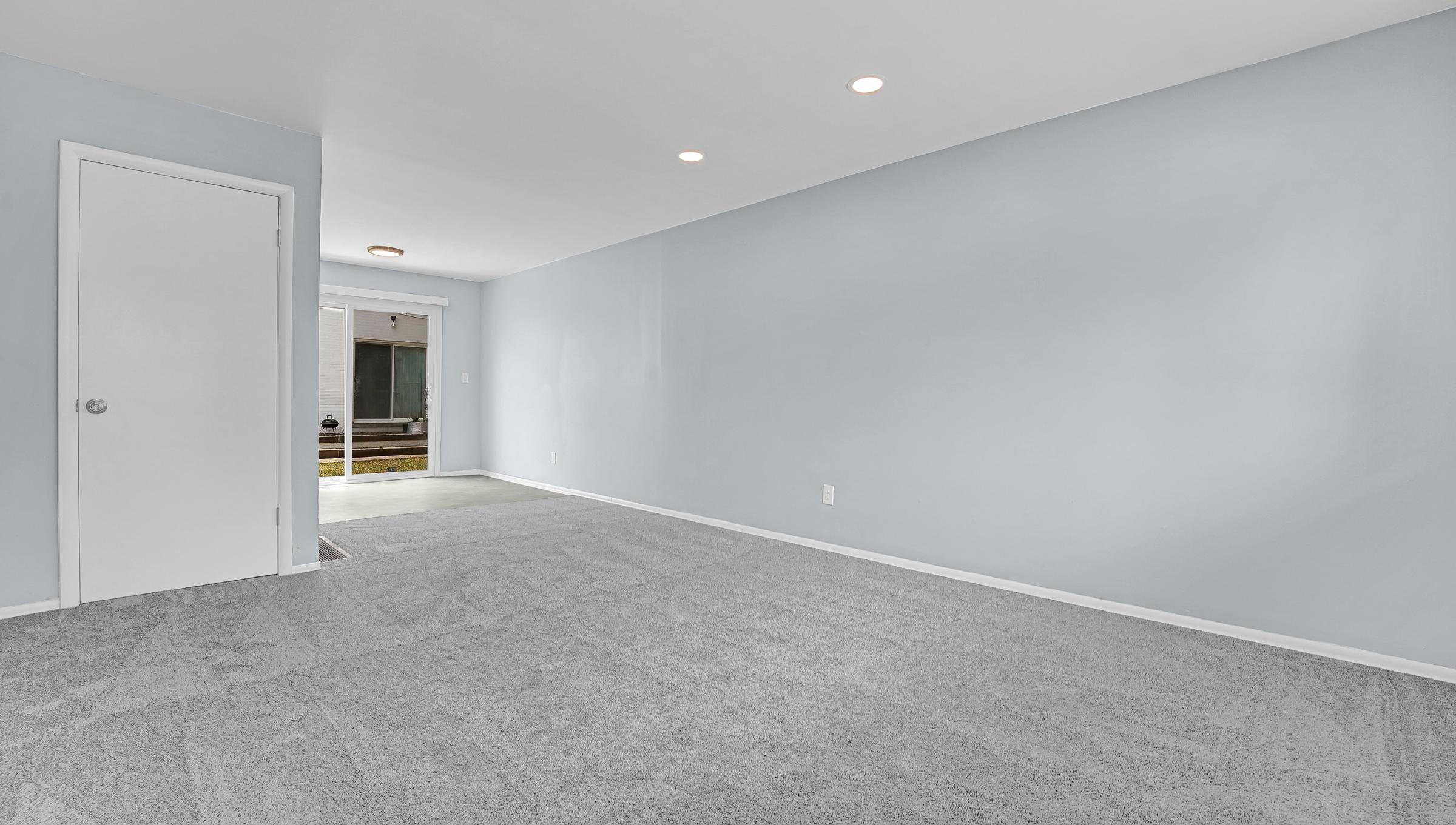 A spacious, empty room with light blue walls and grey carpet. A doorway leads to another area, illuminated by soft overhead lighting. The room is uncluttered, showcasing its potential for various uses or furniture arrangements. Natural light is visible from the outside through the doorway.