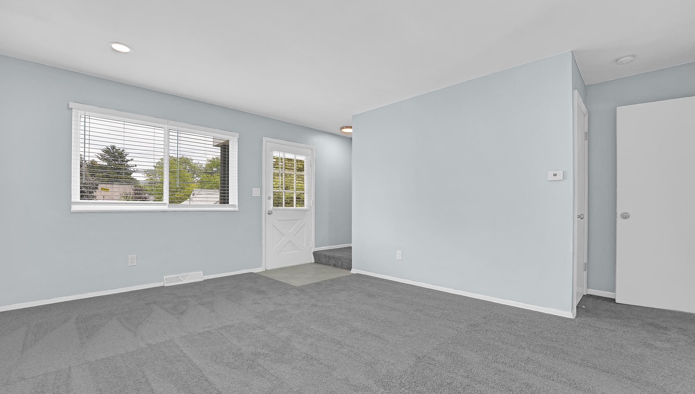 A bright, airy room with light blue walls and gray carpet. It features a white door leading outside, a window with blinds allowing natural light, and a staircase visible in the background. The space is uncluttered and has a minimalist design.