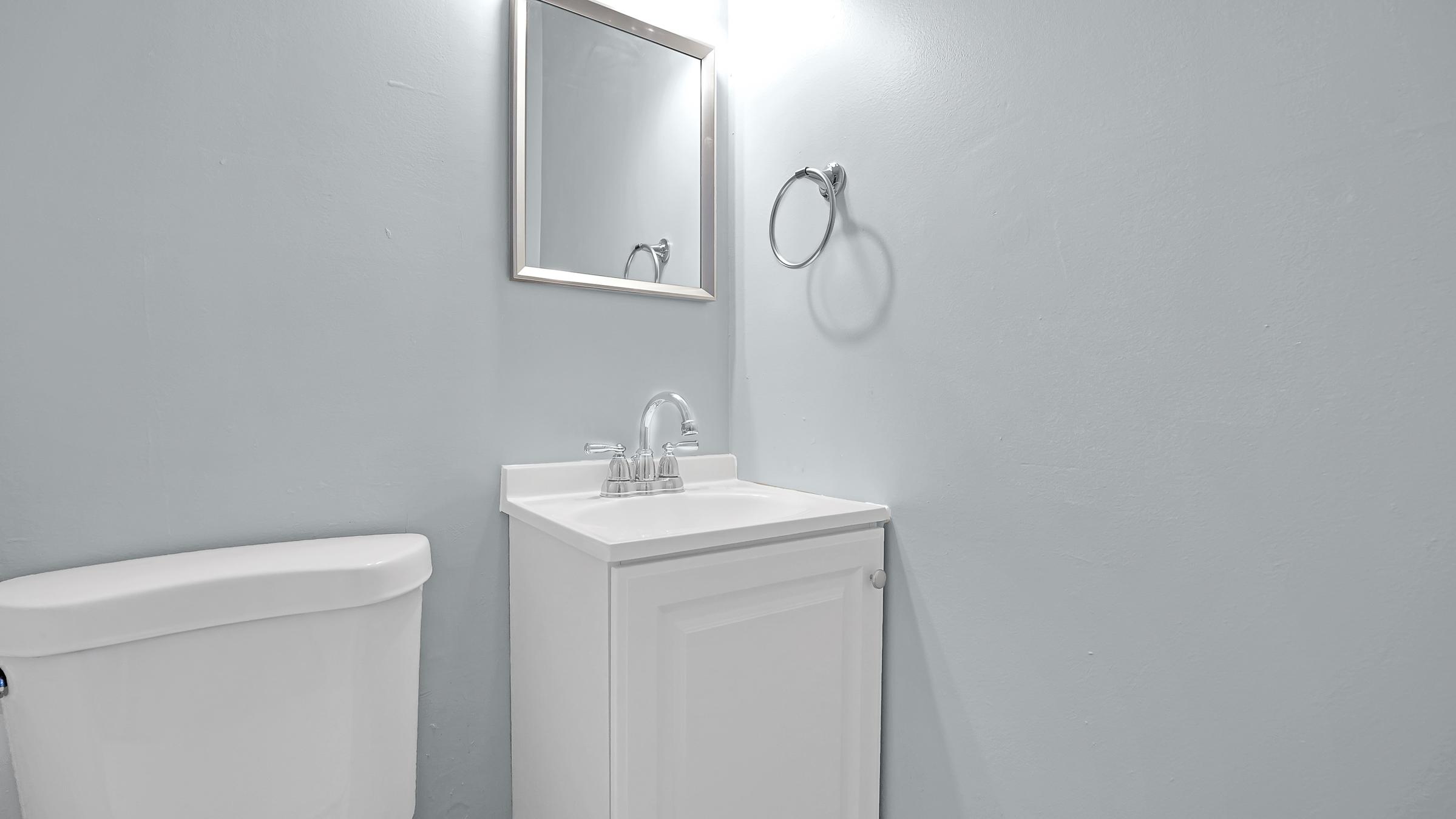 A clean bathroom with light blue walls, featuring a small sink cabinet with a mirror above it. To the left, there is a white toilet. The overall appearance is modern and minimalistic, with good lighting enhancing the serene atmosphere.