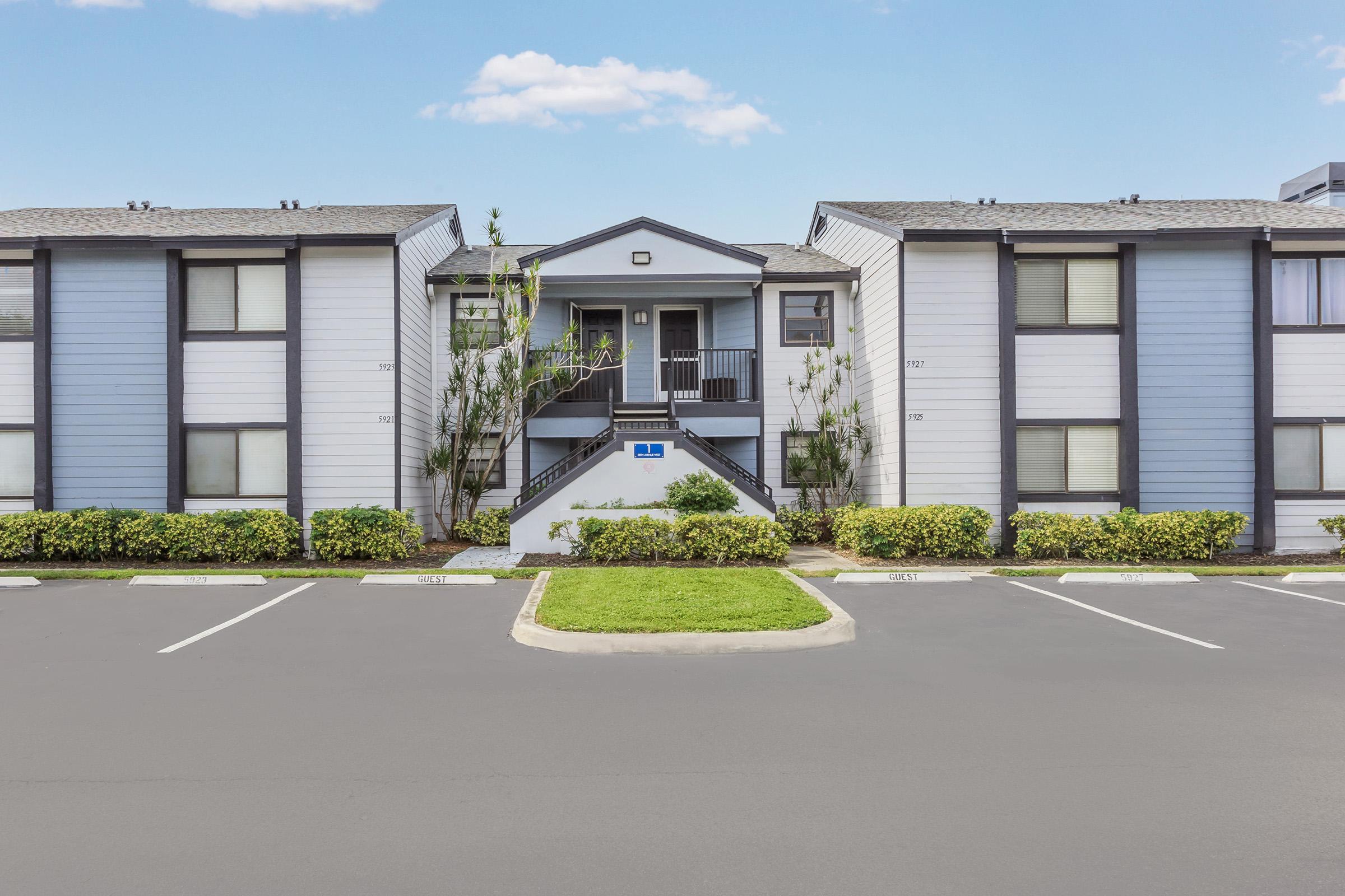 A two-story apartment building with a central entrance surrounded by neatly landscaped bushes and grass. Parking spaces are visible in front of the building, and the sky is clear with a few clouds. The exterior features a combination of light blue and white siding.