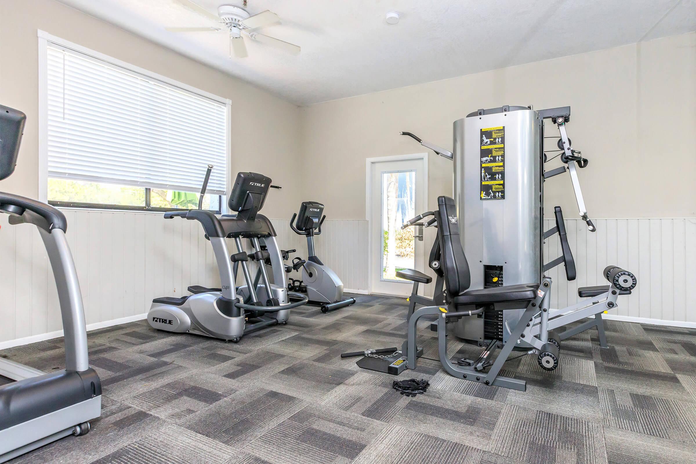 A well-lit gym interior featuring exercise equipment, including an elliptical machine, a stationary bike, and a multi-station weight machine. Large windows allow natural light, and the floor is covered with gray carpet tiles in a checkered pattern. A ceiling fan is visible above.
