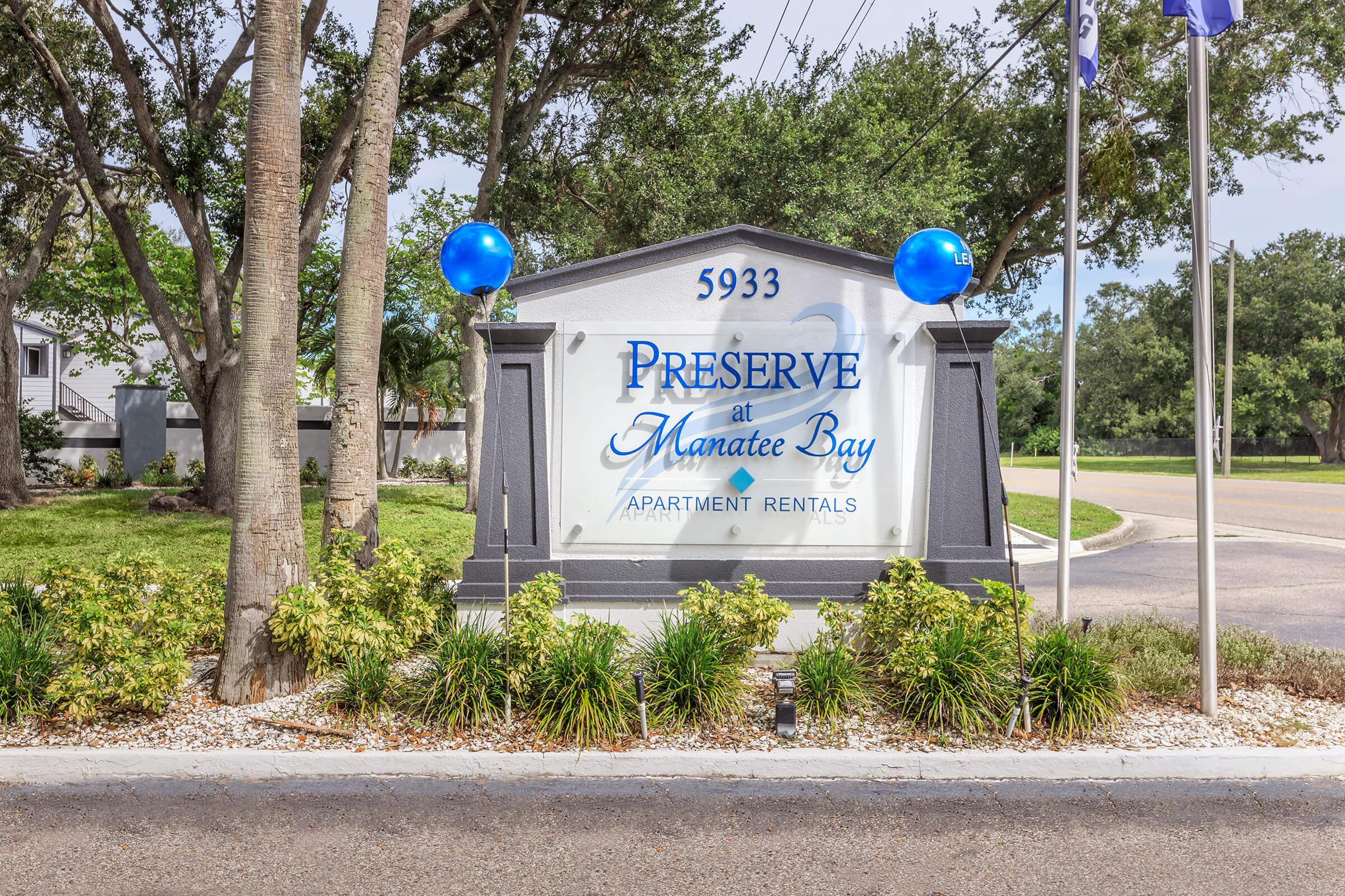 Sign for "Preserve at Manatee Bay" apartment rentals located at 5933, featuring a decorative design with blue balloons and surrounded by greenery.