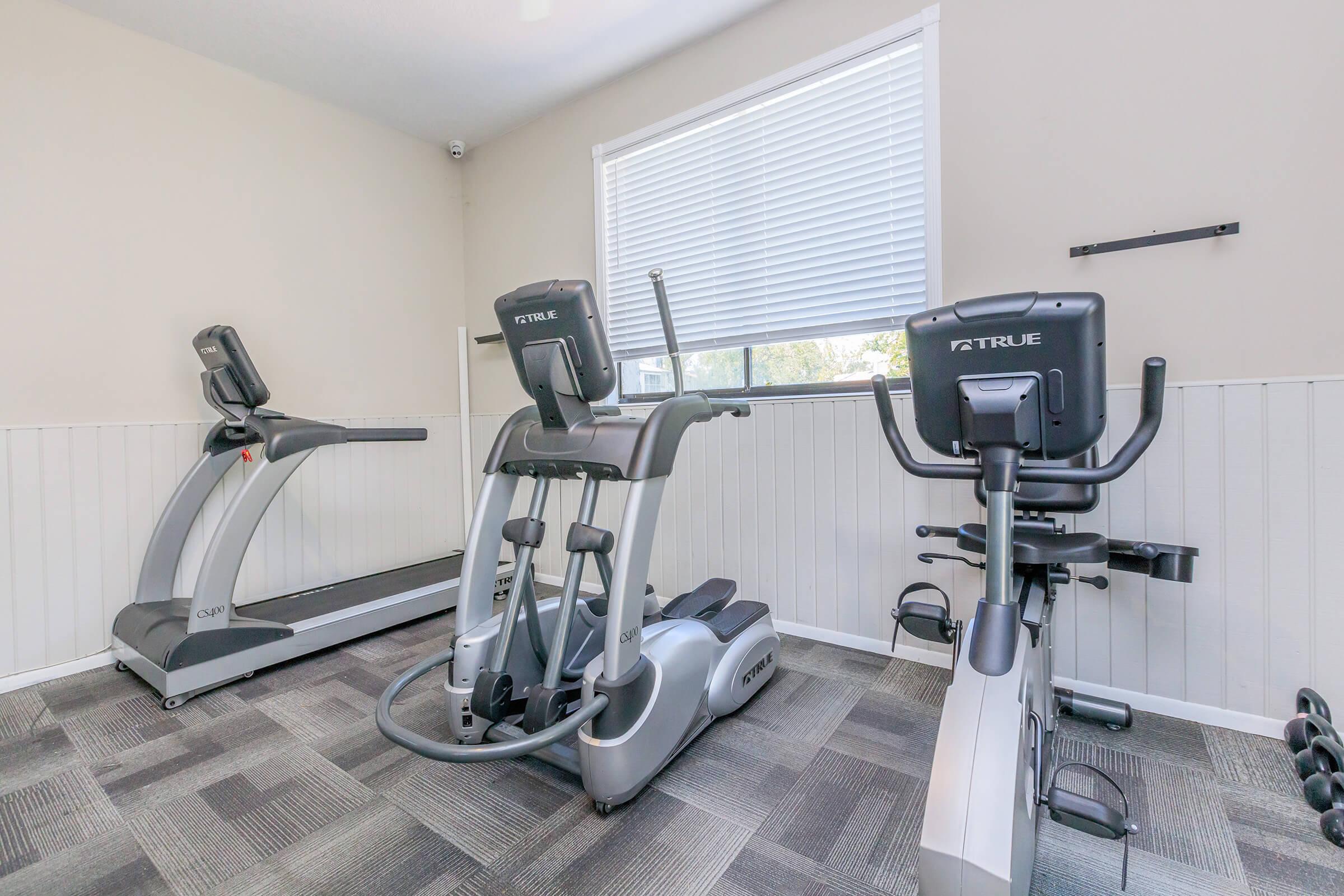 A well-lit fitness room featuring a treadmill, an elliptical machine, and a stationary bike. The space has light-colored walls and a window with blinds, providing a clean and modern atmosphere for exercise. The flooring is carpeted with a geometric pattern.