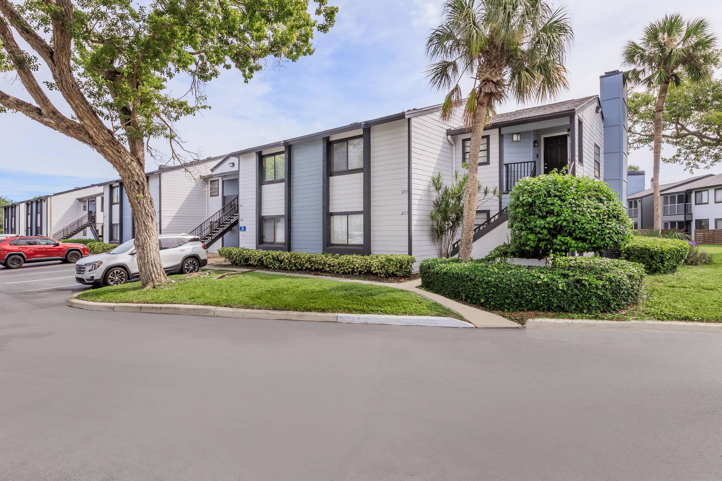 A view of a modern apartment complex featuring two-story buildings with light-colored exteriors, green lawns, and palm trees. Vehicles are parked near the entrance, and the pathway leads to the units. The sky is partly cloudy, suggesting a pleasant day.