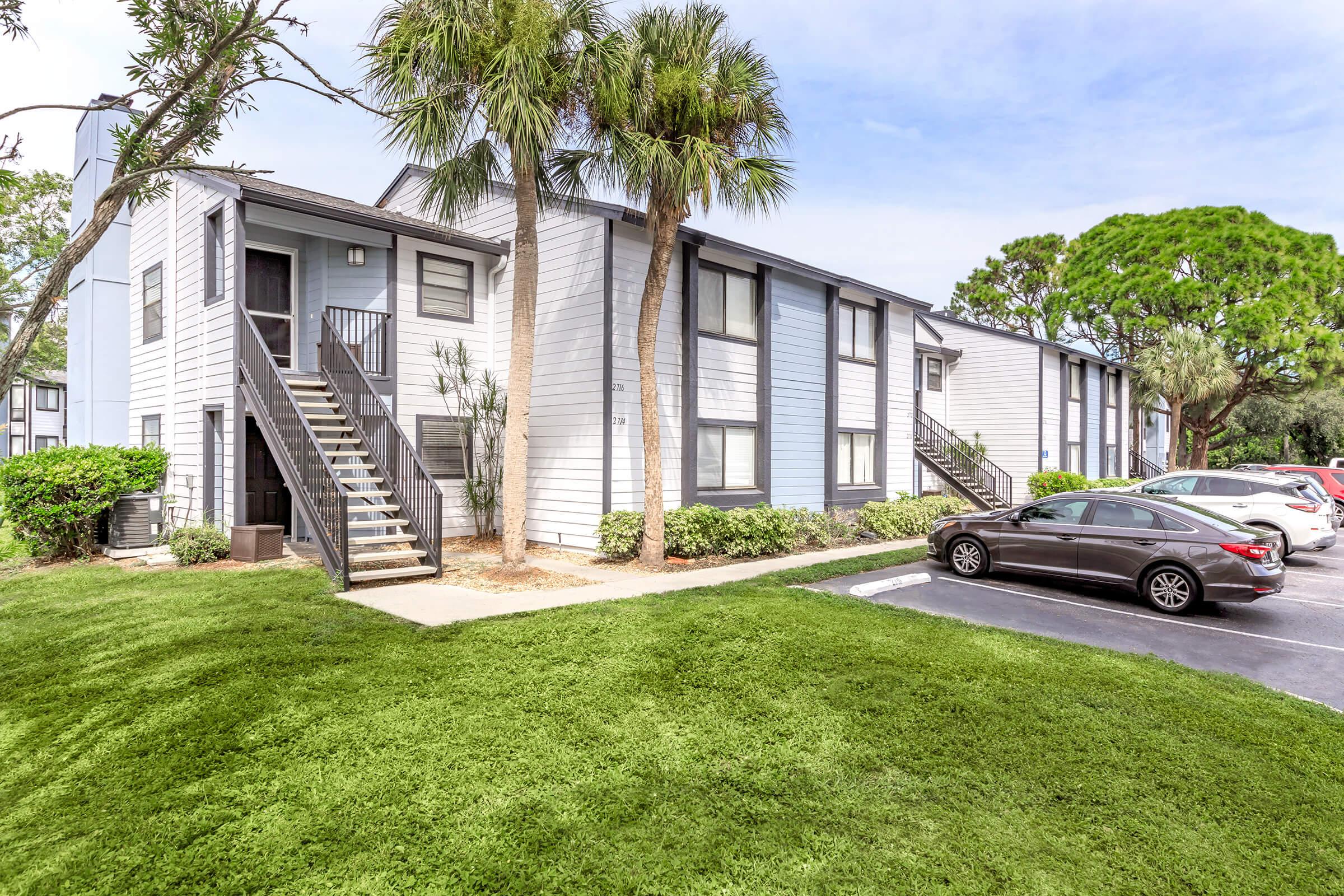 Two-story apartment building with light blue and white siding. Palm trees and green shrubs surround the entrance, which has stairs leading to the upper level. A grassy area in front and several parked cars in a blacktop parking lot. A bright sky enhances the sunny atmosphere.