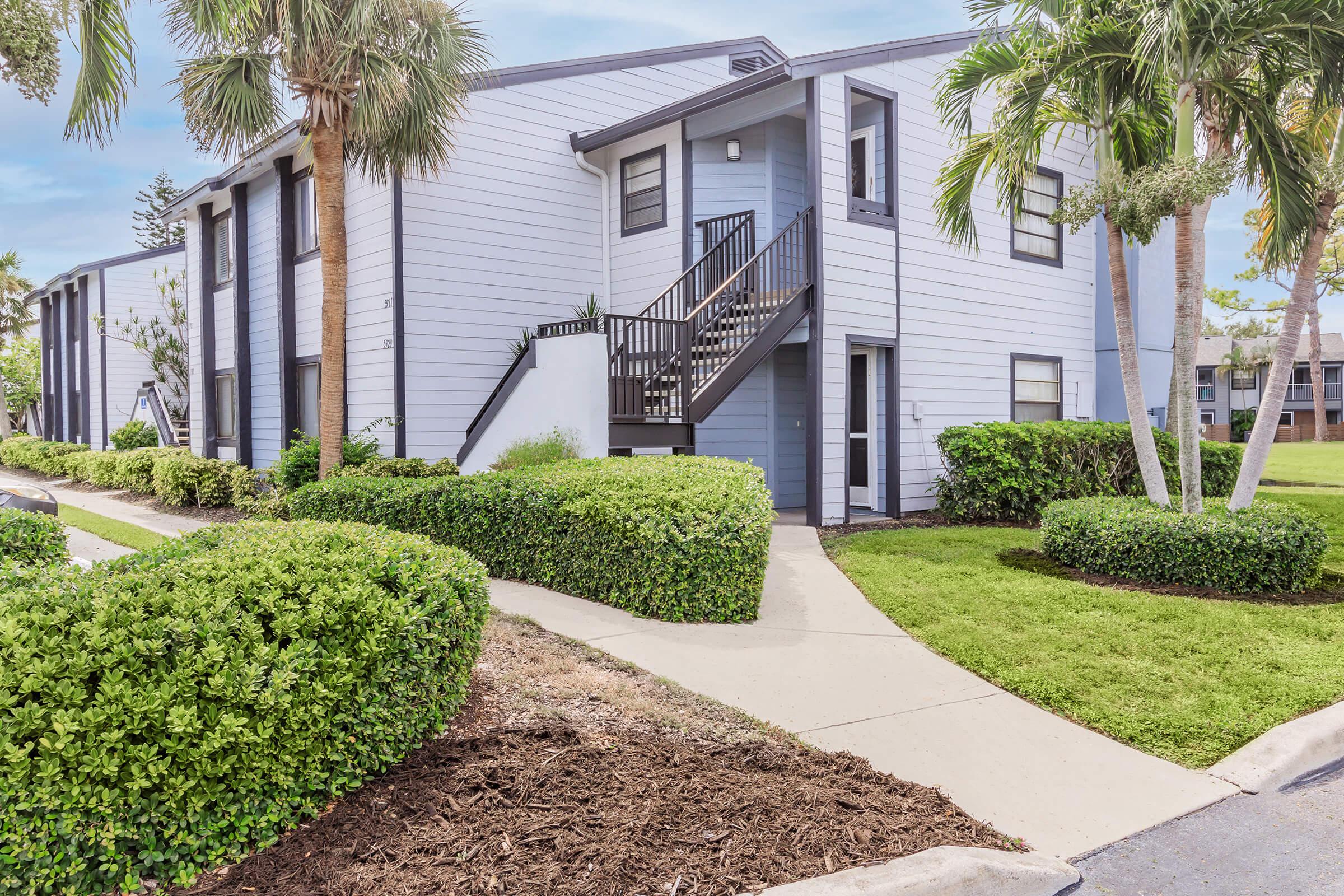A modern two-story apartment building with light blue siding, featuring a set of black stairs leading to the second floor. Well-maintained landscaping includes lush green bushes and palm trees, with a paved walkway leading to the entrance. The scene is bright and inviting, surrounded by a manicured lawn.