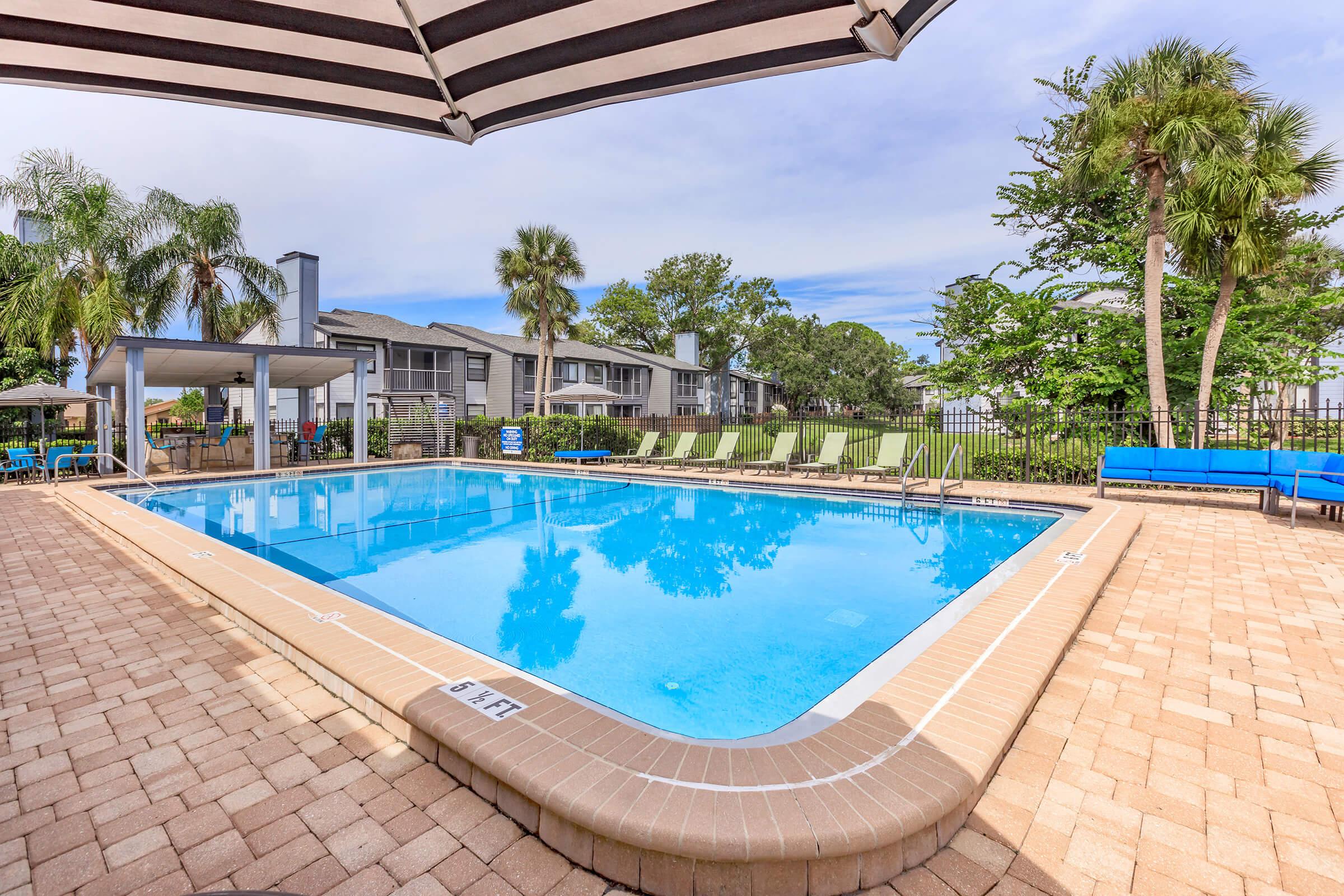 A clear swimming pool surrounded by a patio with lounge chairs. Palm trees and lush greenery are visible in the background, along with a shaded seating area under a striped canopy. The pool area is well-maintained, creating a relaxing outdoor space.