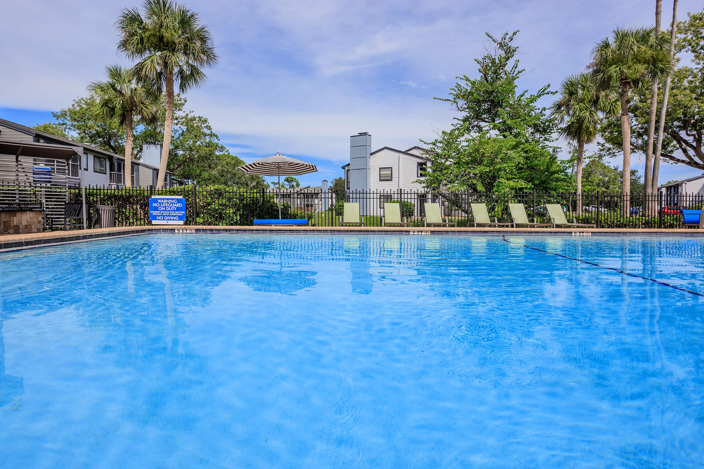 Clear blue swimming pool with lounge chairs and a shaded umbrella in a residential area. Lush greenery and trees line the poolside, with a light-colored building visible in the background under a partly cloudy sky. The scene suggests a relaxing outdoor environment.