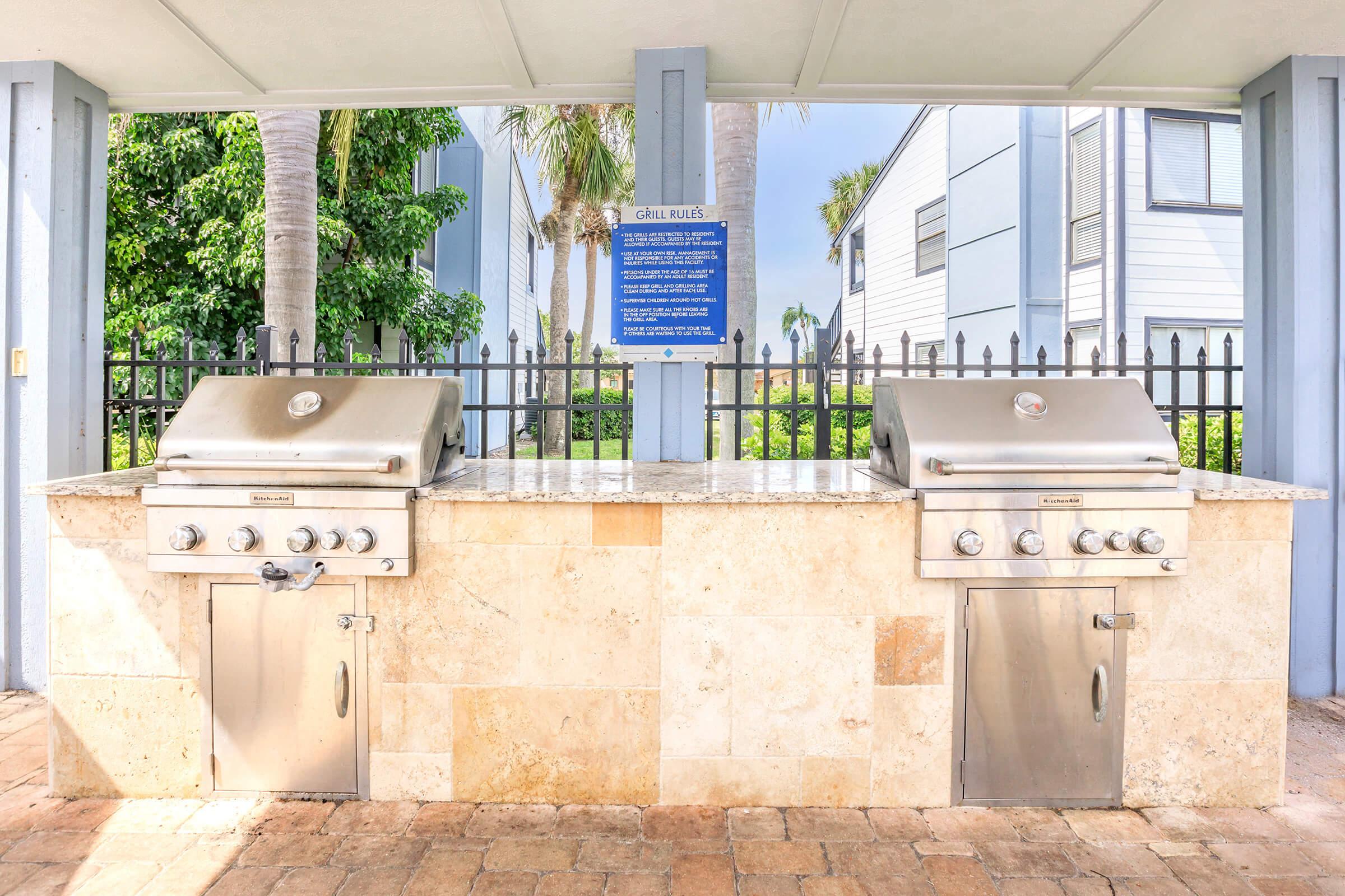 An outdoor grilling area featuring two stainless steel grills on a stone countertop, surrounded by palm trees and a blue sign outlining grilling rules. The space is shaded and includes a fence in the background, with nearby residential buildings visible.