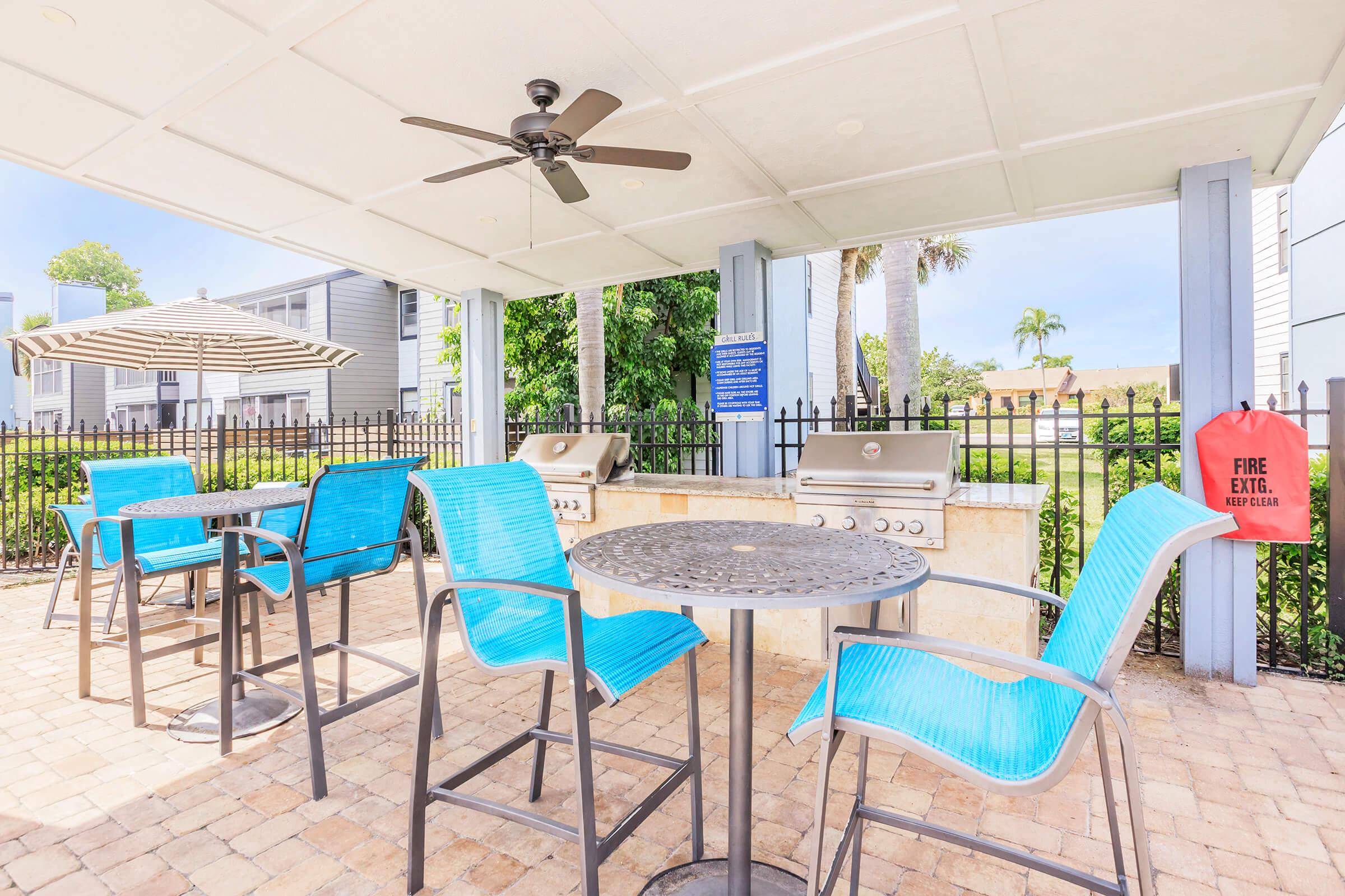 An outdoor barbecue area featuring two grilling stations, a round table with four blue chairs, and a shaded umbrella. The space is surrounded by green landscaping and includes a fire extinguisher mounted on the wall.